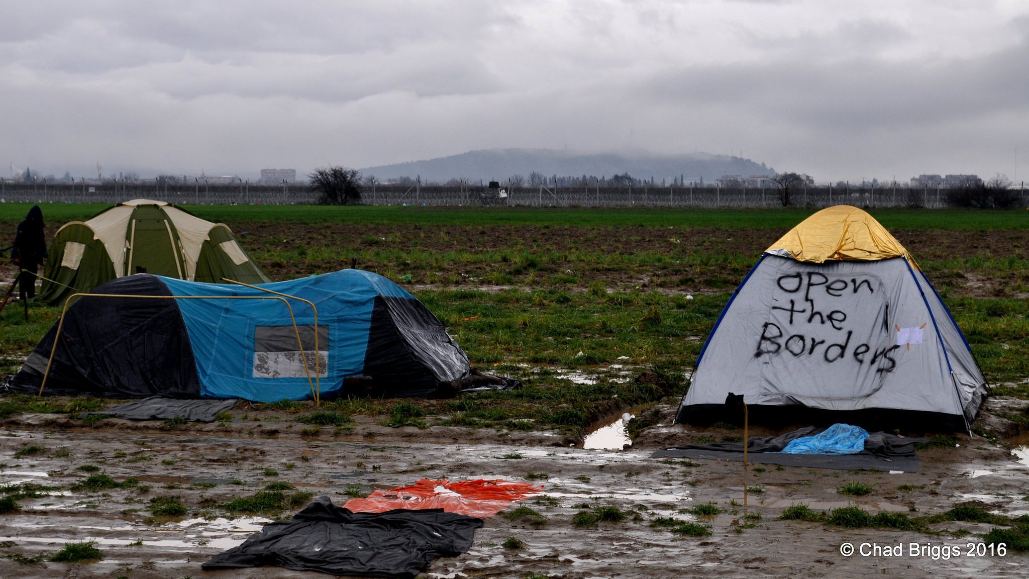L'image montre un campement de tentes installé dans un terrain boueux. On aperçoit plusieurs tentes, dont l'une a un message écrit : "Open the Borders". Le paysage environnant est composé de champs et d'une arrière-plan nuageux, ce qui crée une atmosphère morose. Le sol est détrempé, indiquant probablement des conditions météorologiques difficiles.