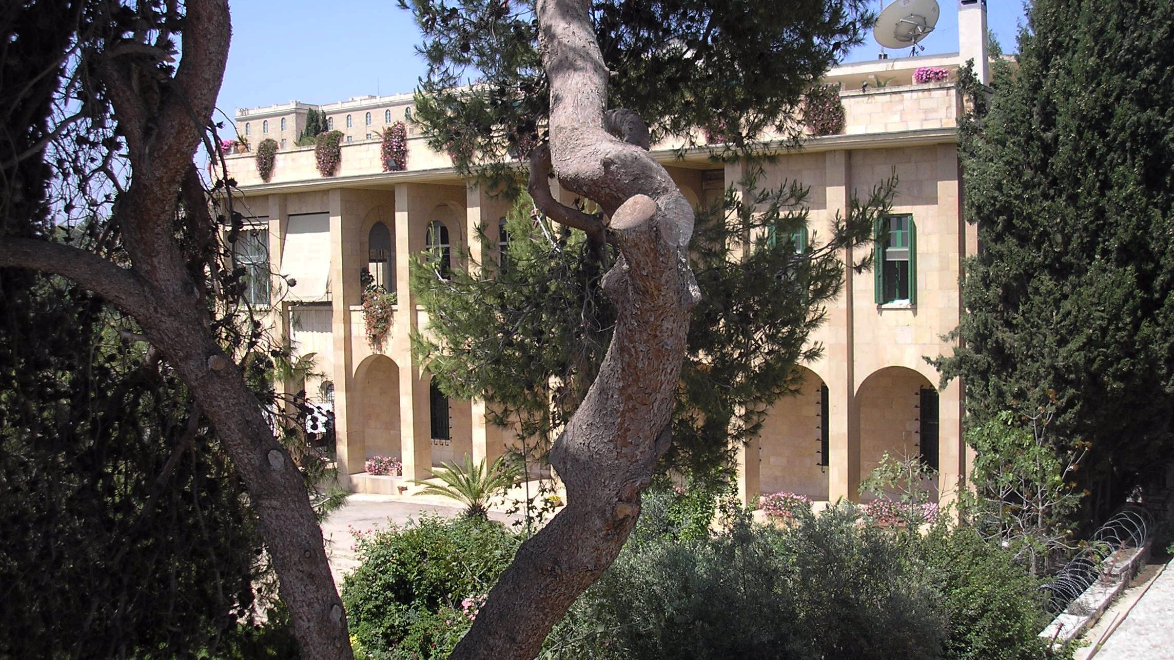 L'image montre un bâtiment en pierre, typique de l'architecture méditerranéenne, avec plusieurs arches et fenêtres. Il est entouré de verdure, avec des arbres et des plantes bien entretenues. Au fond, on remarque un satellite et un ciel bleu clair, ce qui suggère un climat ensoleillé. L'ensemble dégage une atmosphère paisible et accueillante.