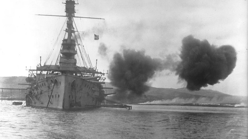 L'image montre un grand navire de guerre, probablement un cuirassé, tirant des armes à feu, ce qui crée des nuages de fumée sur l'eau. Le navire est équipé de plusieurs canons sur ses flancs. En arrière-plan, on aperçoit des collines ou des montagnes, ainsi qu'une éventuelle base maritime ou un autre type d'installations côtières. L'atmosphère est dynamique, accentuée par l'action de tir.