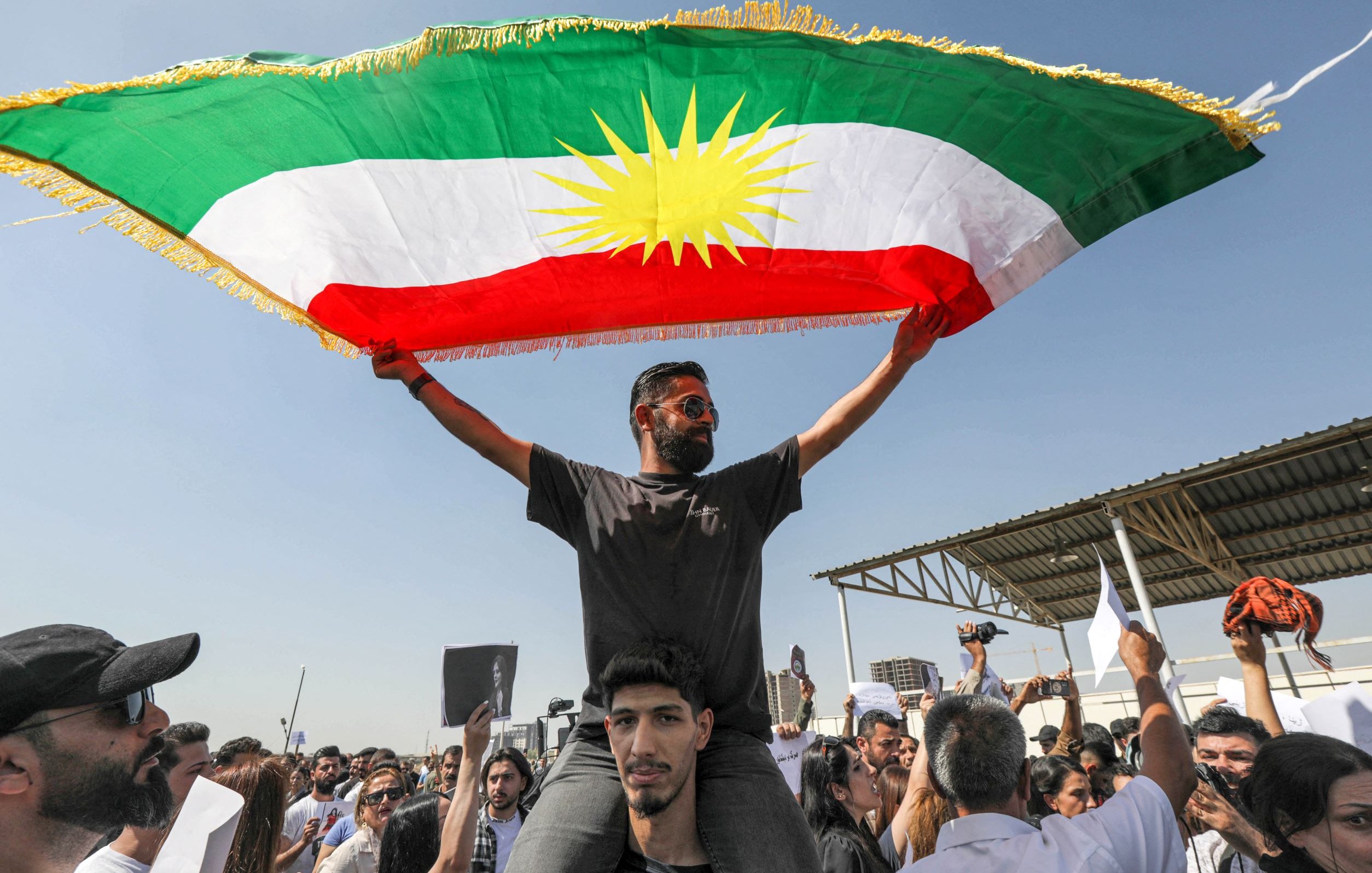 L'image montre un groupe de personnes lors d'une manifestation. Au premier plan, un homme se tient sur les épaules d'un autre, brandissant un grand drapeau kurde, qui est composé de bandes vertes, rouges et blanches avec un soleil jaune au centre. La foule autour d'eux paraît enthousiaste et engagée. Le ciel est clair, ce qui indique une journée ensoleillée. Des pancartes sont également visibles parmi les manifestants, suggérant qu'ils expriment des revendications ou des support pour une cause.