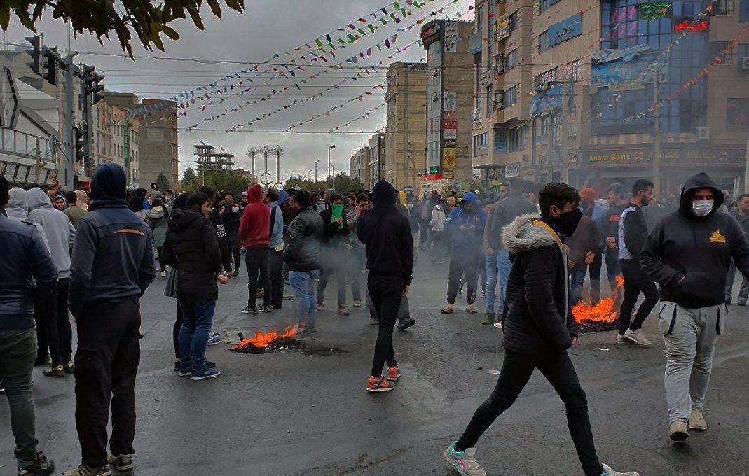 L'image montre une foule rassemblée dans une rue, probablement lors d'une manifestation. Plusieurs personnes portent des vêtements chauds et des masques. On peut observer des petits feux sur la chaussée, entourés de fumée. L'atmosphère semble tendue, avec des bâtiments et des décorations en arrière-plan. Le ciel est nuageux, ajoutant à l'ambiance sombre de la scène.