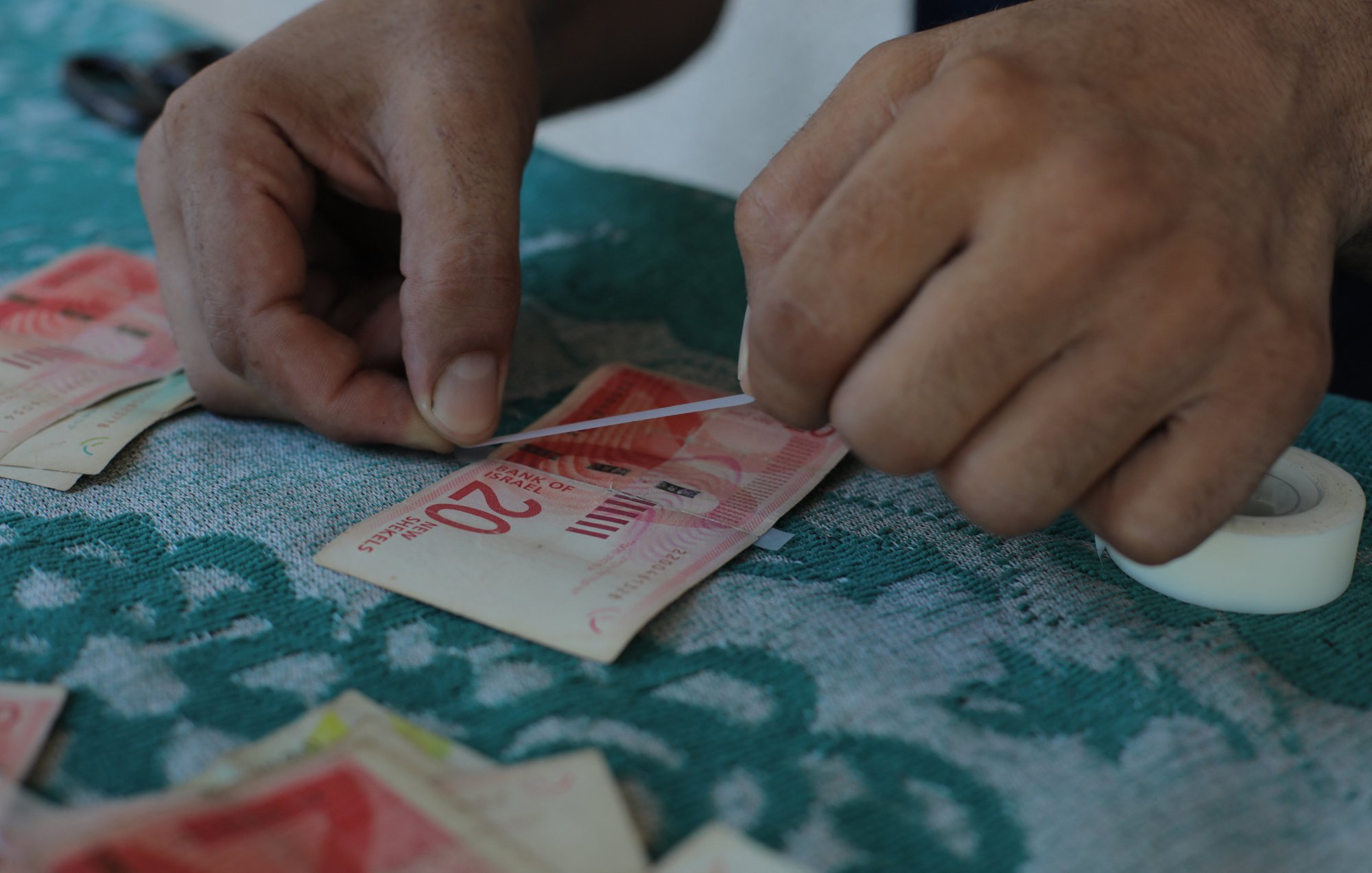 L'image montre des mains qui manipulent des billets de banque. Une personne semble être en train de recoller ou d'ajuster un billet de 20 unités. On peut voir un surface en tissu avec des motifs, ainsi que d'autres billets éparpillés autour. La situation donne une impression de travail minutieux avec une attention particulière aux détails.