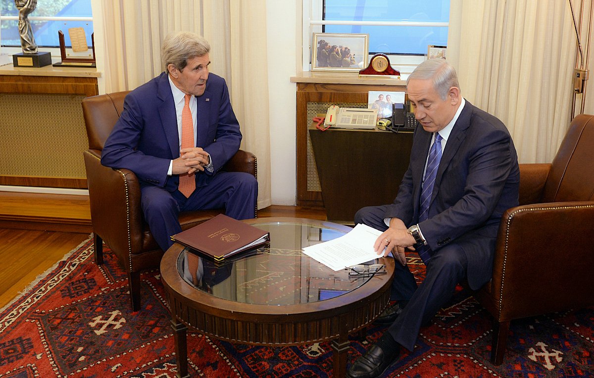 L'image montre deux hommes assis dans une pièce restructurée. L'un est vêtu d'un costume orange et l'autre d'un costume sombre, tous deux semblent être en discussion sérieuse. Ils sont assis autour d'une table basse en bois, avec des documents devant eux. En arrière-plan, on aperçoit une fenêtre et une décoration sobre, suggérant un cadre officiel et professionnel.