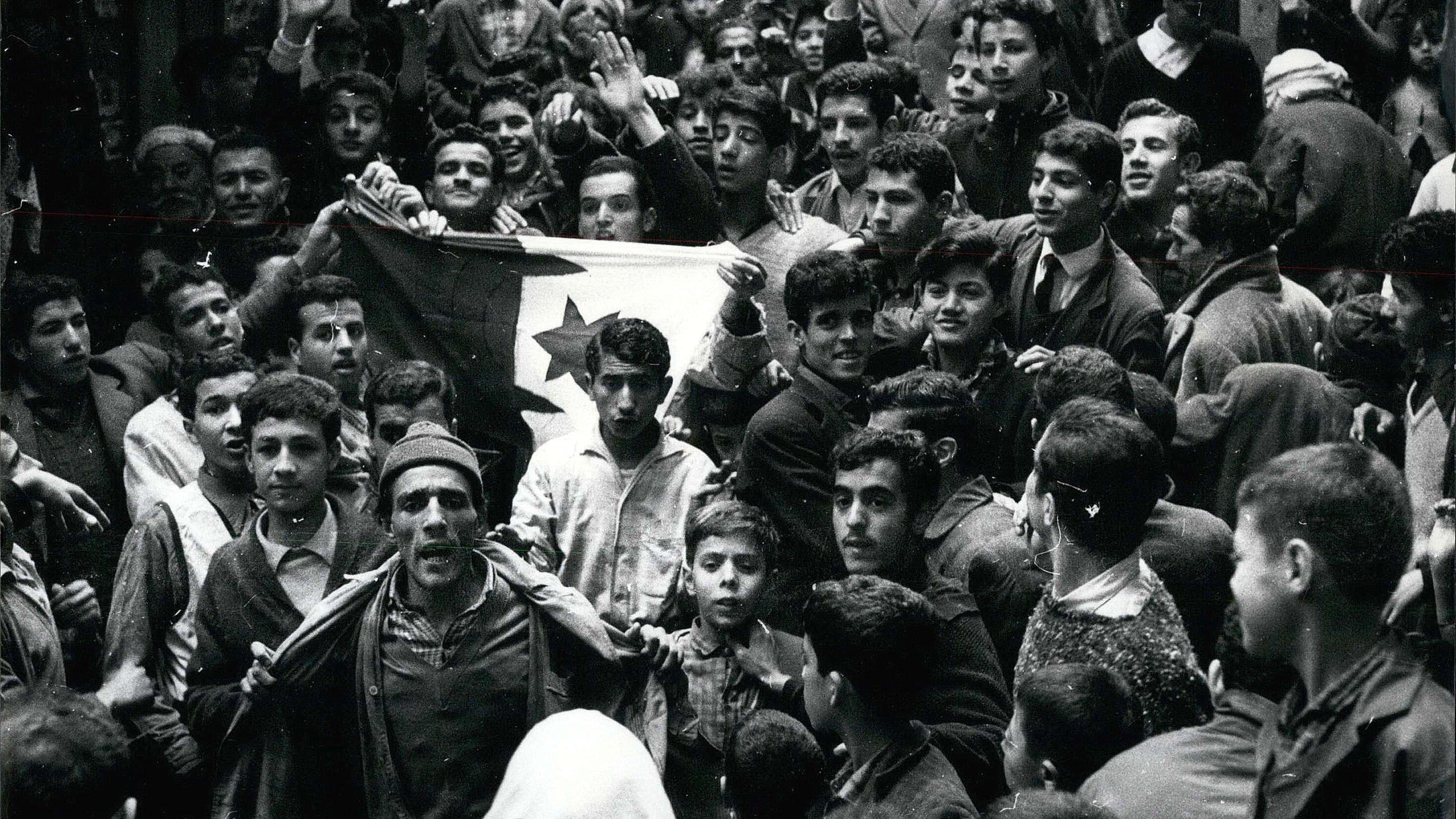L'image montre une foule dense de personnes rassemblées dans une rue. Au premier plan, plusieurs individus sont visibles, dont un homme qui semble porter un drapeau avec un symbole distinctif. Les expressions des visages varient, certains levant les bras en signe de soutien ou d'enthousiasme. On peut observer des jeunes, des adultes et même des enfants parmi la foule. L'atmosphère semble intense et chargée d'émotion, indiquant un moment significatif ou une manifestation. La scène est en noir et blanc, ce qui lui donne un aspect historique.