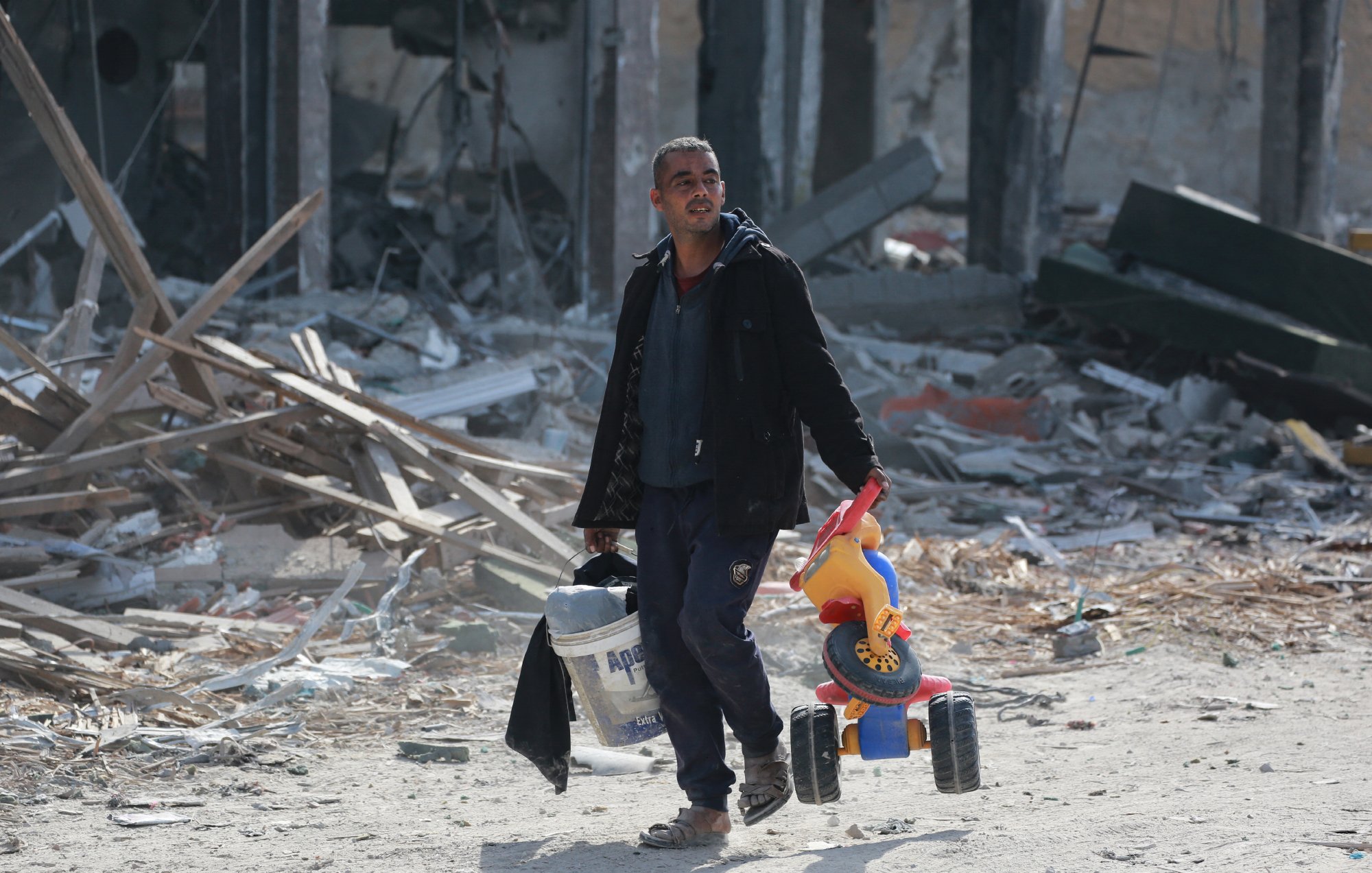 L'image montre un homme marchant dans un paysage dévasté. Il porte un seau de peinture dans sa main gauche et un jouet pour enfant, un petit véhicule à roues, dans sa main droite. L'environnement autour de lui est marqué par des débris et des ruines, suggérant un endroit qui a subi des destructions importantes. L'homme a l'air déterminé malgré la situation difficile.