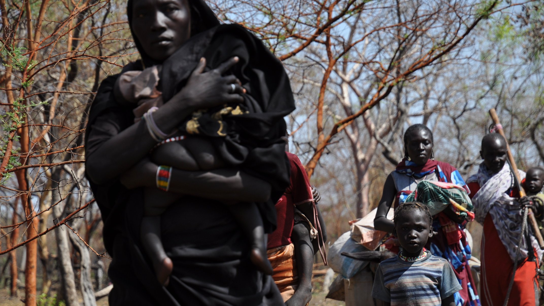 L'image montre un groupe de personnes se déplaçant dans une zone boisée. Au premier plan, une femme porte un enfant sur son dos, tandis qu'à côté d'elle, un petit garçon regarde vers l'avant. D'autres femmes et enfants les suivent, portant des sacs et des objets sur leurs épaules. Leurs vêtements sont colorés, et le décor est constitué d'arbres aux branches dénudées, suggérant un environnement aride. L'atmosphère semble être celle d'un déplacement ou d'une communauté en mouvement.