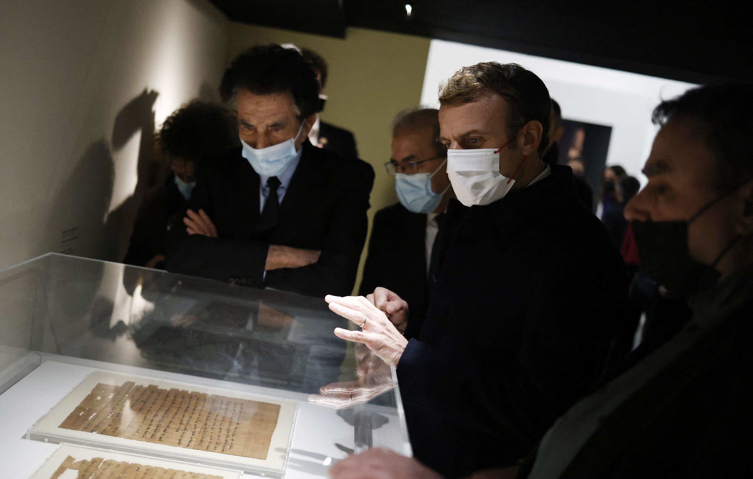 L'image montre un groupe de personnes, dont une figure centrale qui semble être un homme en train d'observer des documents exposés dans une vitrine. Ils portent tous des masques, probablement en raison de mesures sanitaires. L'environnement est sombre, avec des murs clairs, et il y a un intérêt évident pour les objets présentés. Les autres personnes dans le cadre semblent également attentives et engagées dans la discussion.