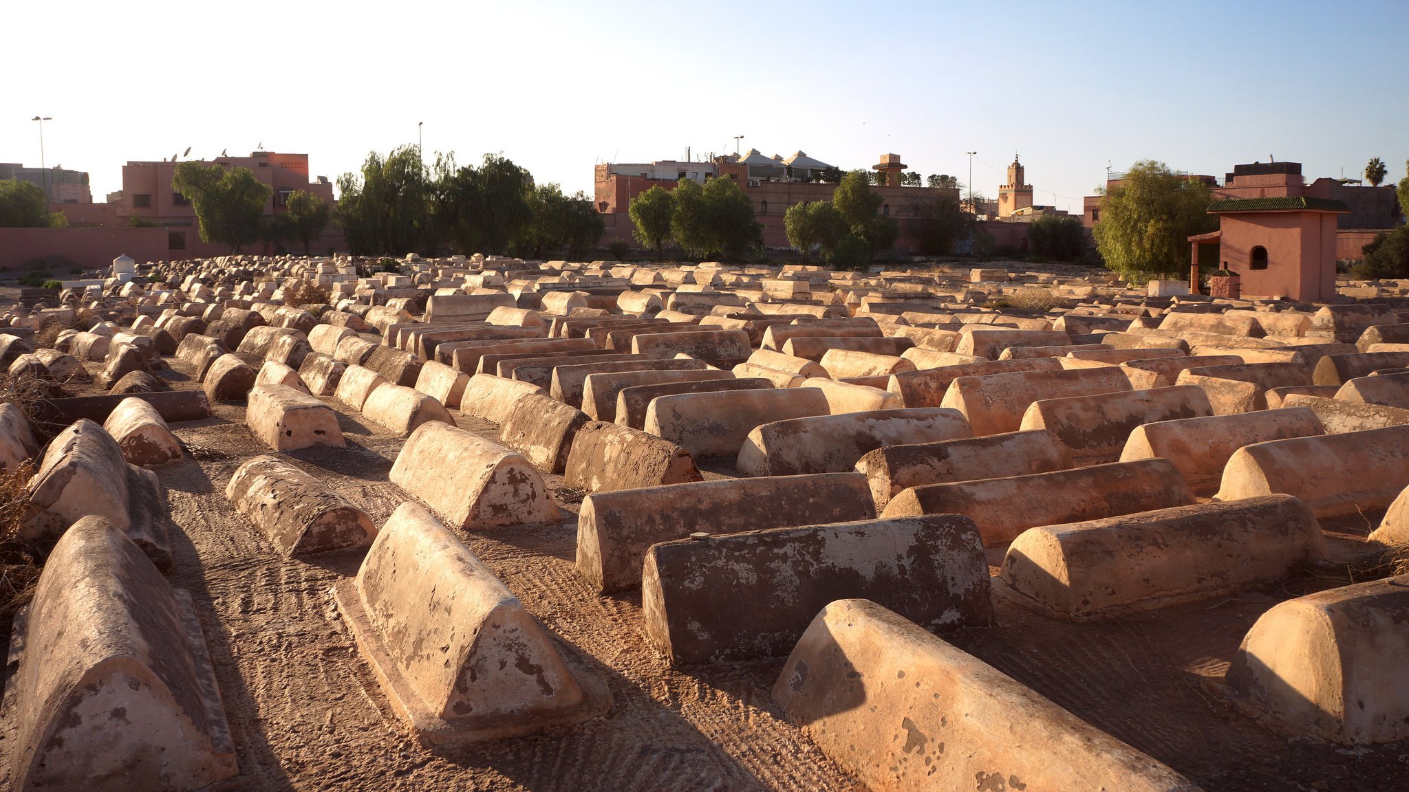 L'image montre un cimetière où l'on peut voir des tombes en forme de tombeaux, étalées sur un terrain sec. Les structures sont de couleur beige ou terre cuite et semblent être en béton, avec une surface usée par le temps. En arrière-plan, on aperçoit des bâtiments et des arbres, indiquant que le cimetière est situé près d'une ville. La lumière du soleil crée une atmosphère calme et paisible, tandis que le ciel est dégagé, laissant entrevoir un beau jour.