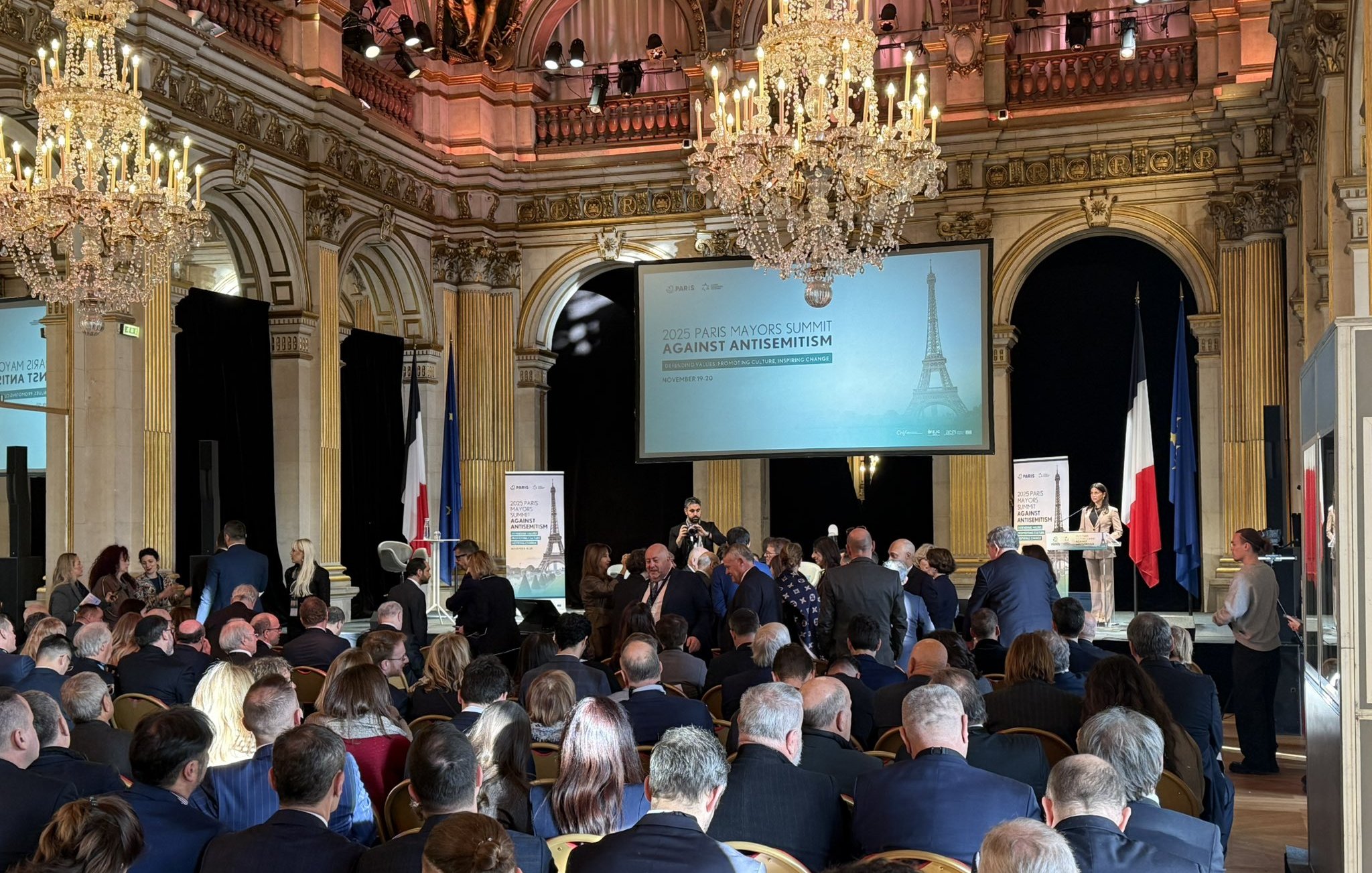 Grand hall avec chandeliers, foule réunie autour d'un discours contre l'antisémitisme.