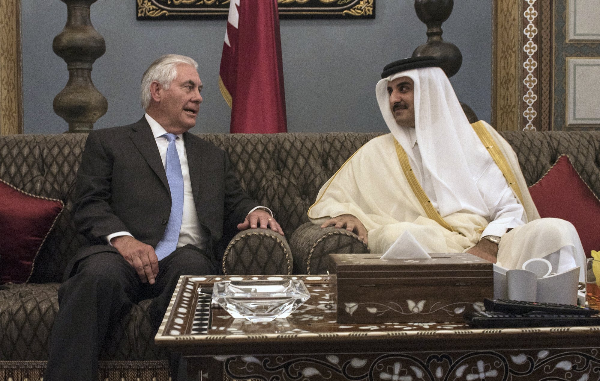 L'image montre deux hommes assis dans un cadre élégant. L'un d'eux porte un costume noir avec une cravate claire, tandis que l'autre est habillé d'une tunique traditionnelle blanche et d'un manteau beige. Ils semblent engagés dans une conversation conviviale, entourés de décorations raffinées, y compris des coussins rouges et des ornements sur la table. L'atmosphère est de camaraderie et de diplomatie.