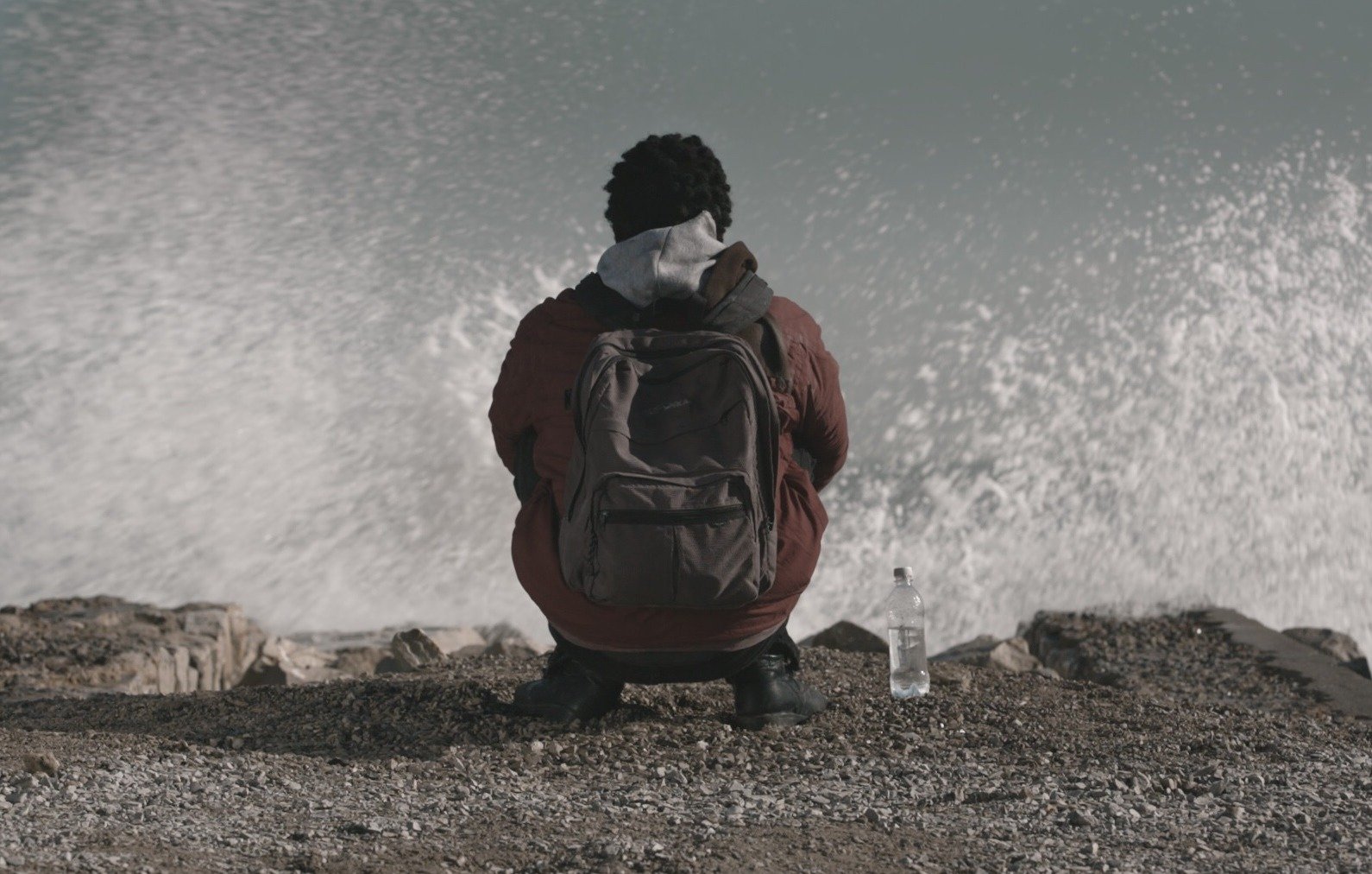 L'image montre une personne assise sur un rivage, tournant le dos à la caméra. Elle porte une veste et un sac à dos. À côté d'elle, il y a une bouteille d'eau. On peut voir des vagues qui se brisent sur les rochers, créant un effet de mouvement dans l'eau. L'atmosphère semble calme mais dynamique en raison du mouvement des vagues.