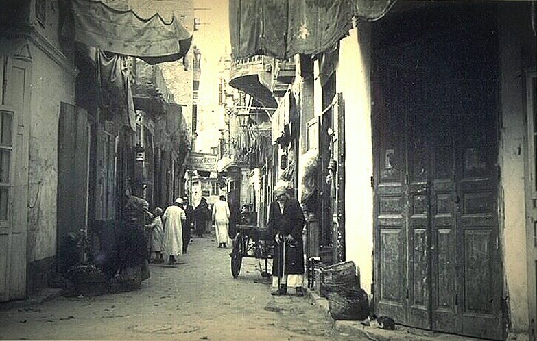 L'image montre une ruelle étroite d'une ville ancienne, probablement dans un pays arabe. Les murs des bâtiments sont ornés de tentures et de draps qui ajoutent une ambiance colorée. On peut voir des habitants, dont certains portent des vêtements traditionnels, déambulant dans la rue. Un homme semble se tenir près d'une charrette, tandis que d'autres personnes se déplacent en arrière-plan. Les portes en bois, typiques, ajoutent un charme ancien à la scène, qui évoque une atmosphère de vie quotidienne dans un vieux quartier.