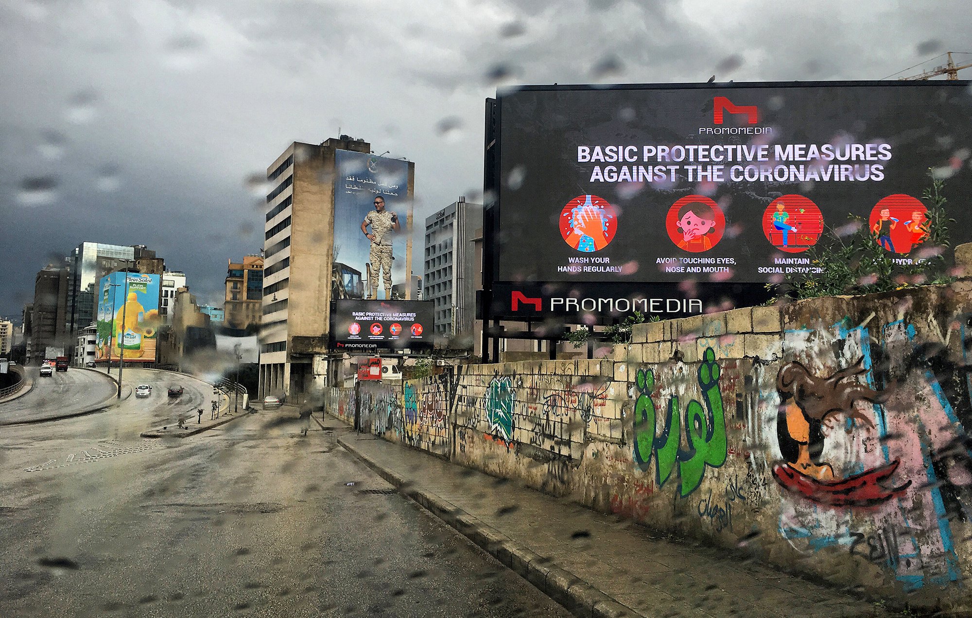 L'image montre une scène urbaine sous la pluie avec des bâtiments modernes à l'arrière-plan. On peut voir un panneau d'affichage numérique qui présente des mesures de protection contre le coronavirus, avec des icônes illustratives. Le mur à gauche est couvert de graffitis colorés, ajoutant une touche artistique à l'environnement. L'atmosphère est plutôt sombre en raison du temps nuageux et pluvieux.
