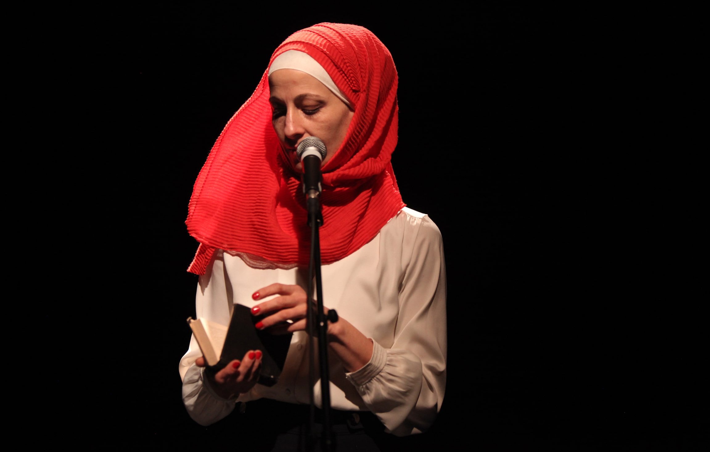 L'image montre une femme debout devant un microphone sur scène. Elle porte un hijab rouge vif et une blouse blanche. Elle tient un petit livre dans ses mains, semblant s'apprêter à lire ou à partager un texte. L'éclairage est sombre, mettant en valeur son visage et l'objet qu'elle tient. L'atmosphère semble intime et concentrée.