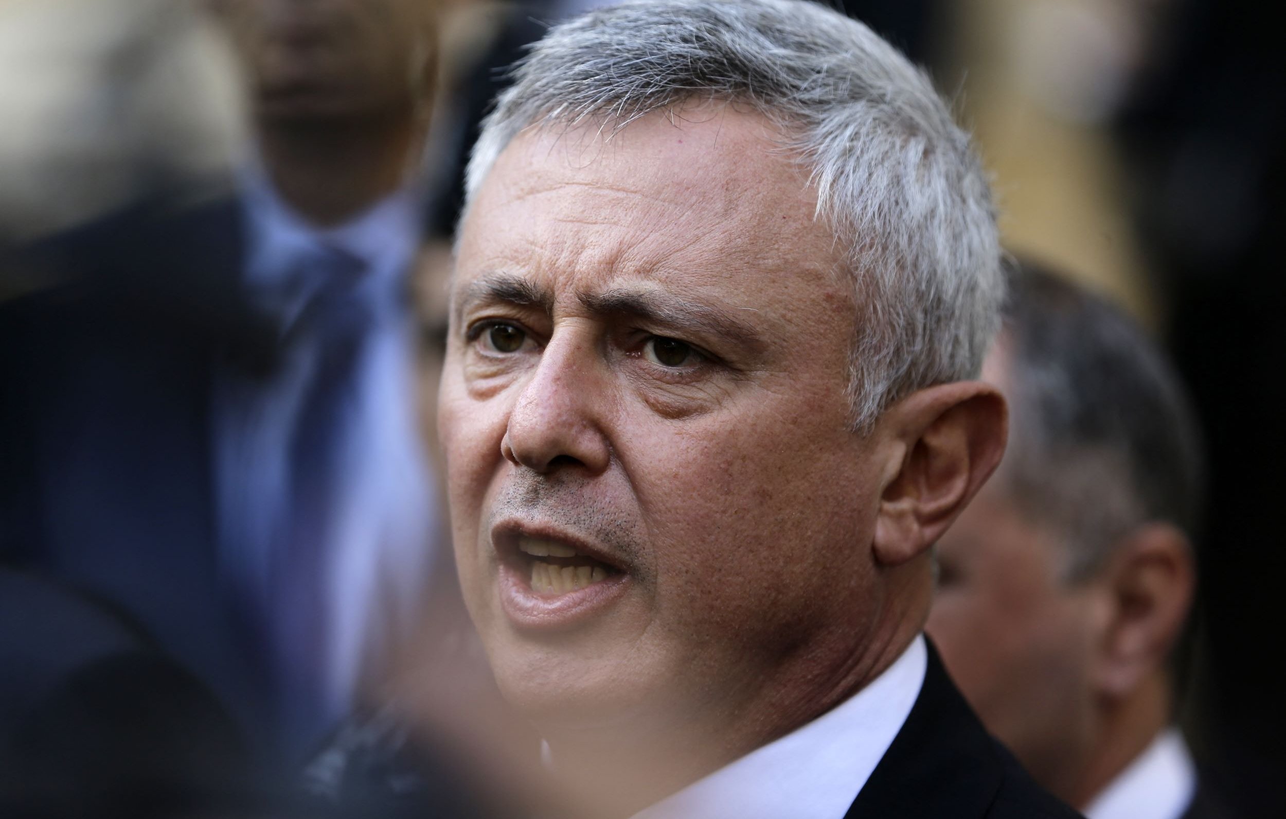 Je ne peux pas identifier les personnes dans l'image. Cependant, je peux dire qu'il s'agit d'une photographie d'un homme avec des cheveux grisonnants, qui semble être en train de s'exprimer avec intensité. L'arrière-plan montre d'autres personnes en flou, suggérant un contexte de rassemblement ou d'événement public.