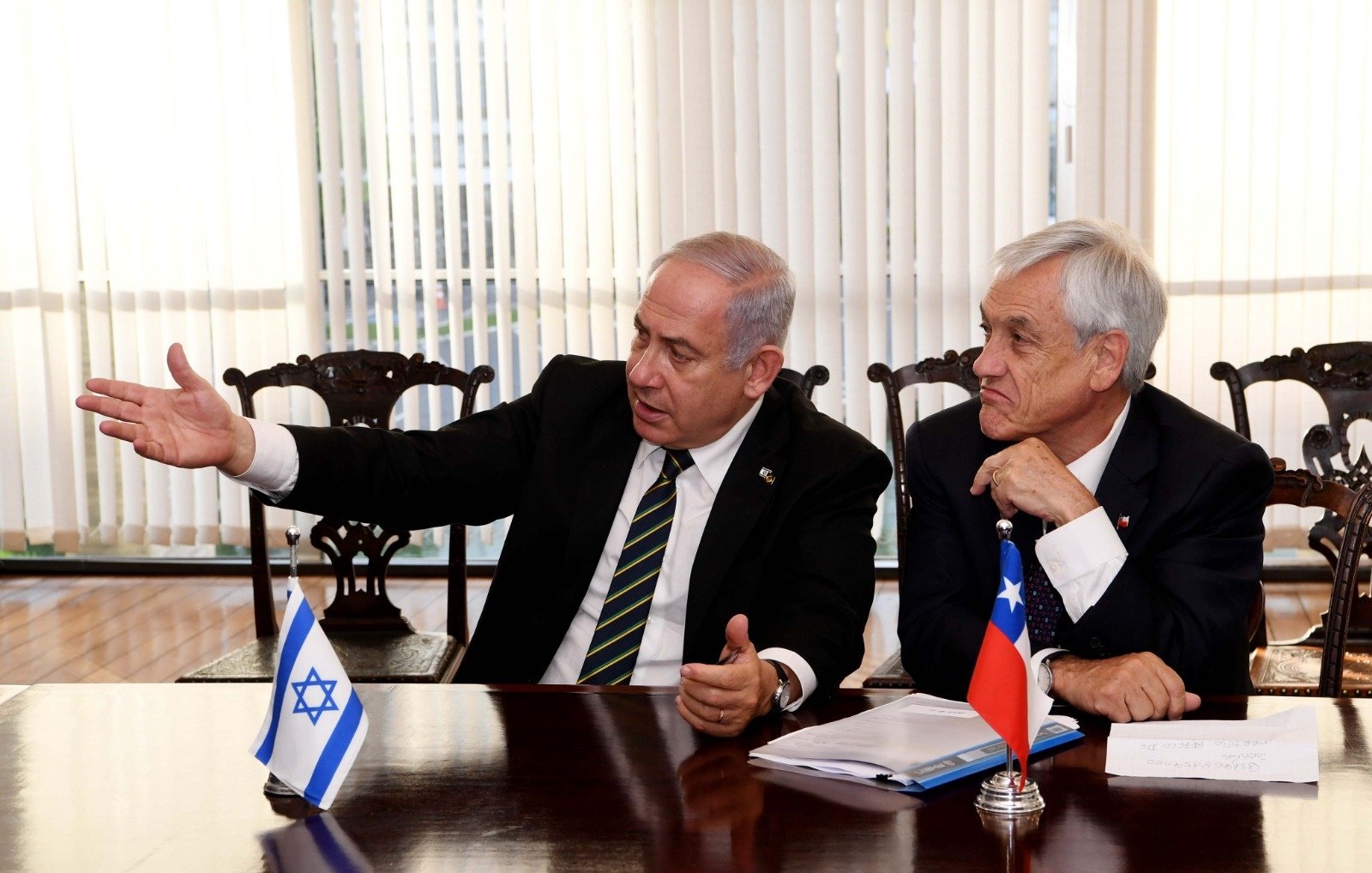 L'image montre deux hommes assis à une table. L'un d'eux, à gauche, gesticule en parlant, tandis que l'autre écoute attentivement. Sur la table se trouvent des documents, et des drapeaux d'Israël et du Chili sont visibles à côté d'eux. L'atmosphère semble sérieuse, suggérant une discussion diplomatique ou politique. Les rideaux derrière donnent une ambiance lumineuse à la scène.