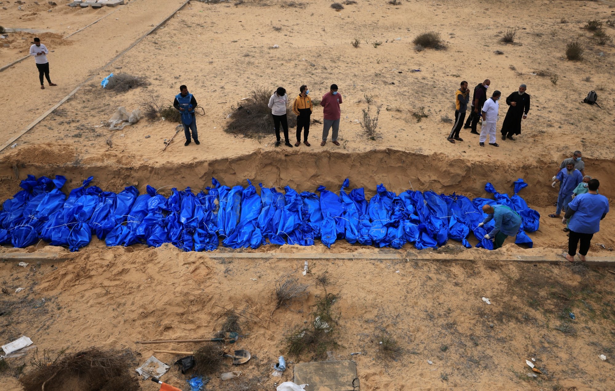 Des corps enveloppés de bleu sont placés dans une fosse, entourés de personnes en deuil.