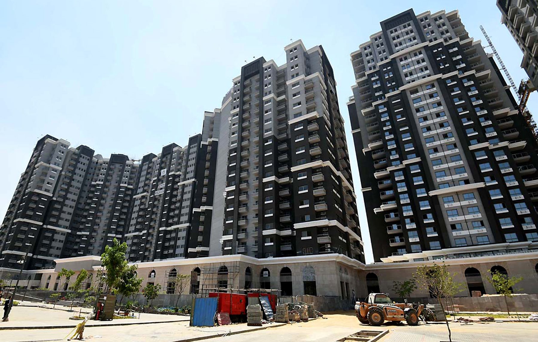 L'image montre un ensemble de grands immeubles résidentiels modernes. Les bâtiments sont principalement de couleur noire et blanche, avec de grandes fenêtres qui laissent entrer la lumière. On peut voir un chantier en cours au premier plan, avec des véhicules de construction et des matériaux dispersés. Il y a aussi quelques arbres et des espaces verts, suggérant un aménagement paysager en développement. Le ciel est dégagé et ensoleillé.