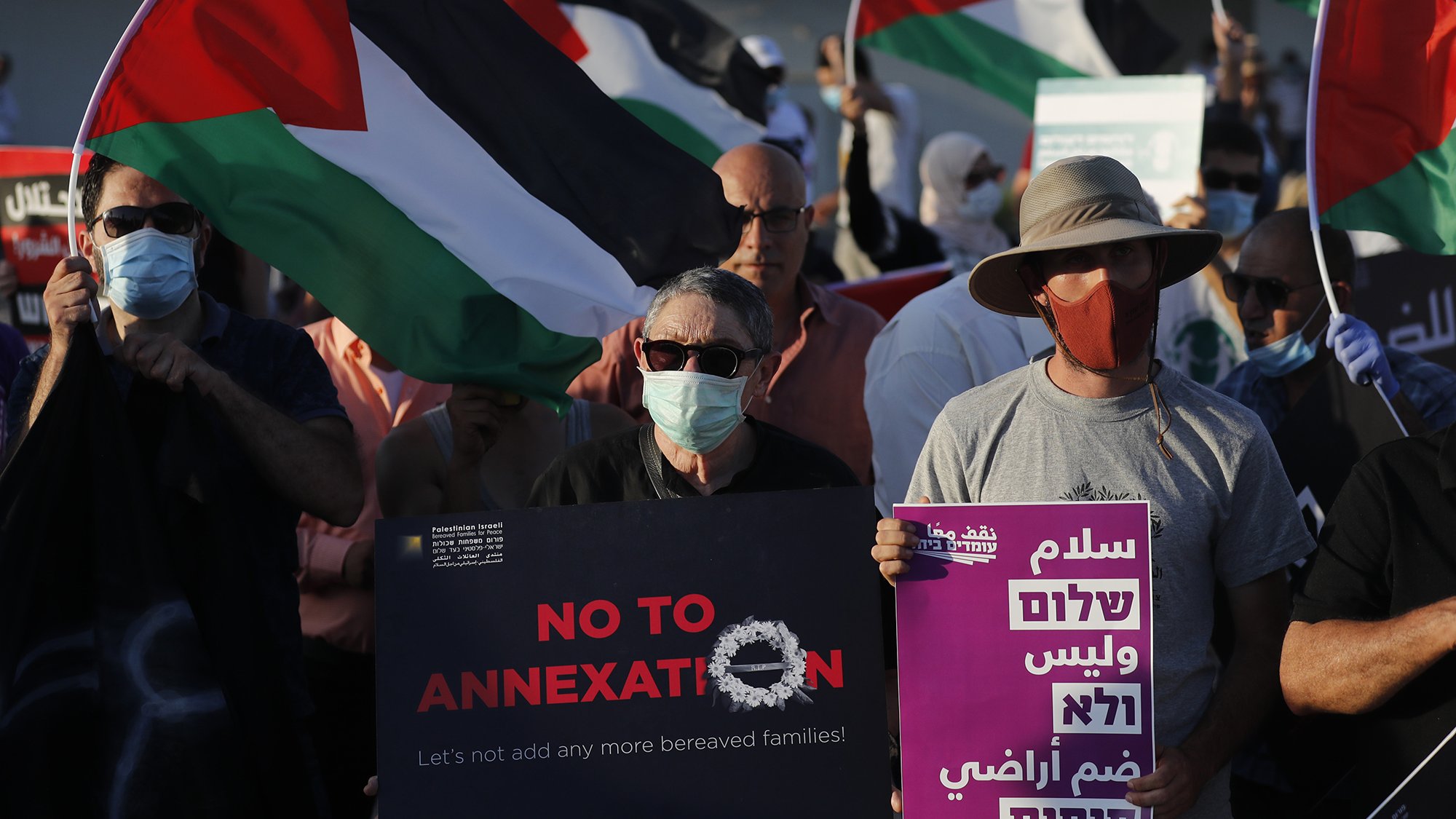 L'image montre une manifestation avec des participants portant des masques. Plusieurs personnes tiennent des drapeaux palestiniens. Des pancartes sont visibles, certaines affichant des messages tels que "NO TO ANNEXATION" (Non à l'annexion) et d'autres slogans en arabe et en hébreu. L'ambiance semble être celle d'une mobilisation pour une cause sociale ou politique, avec un engagement visible des manifestants.