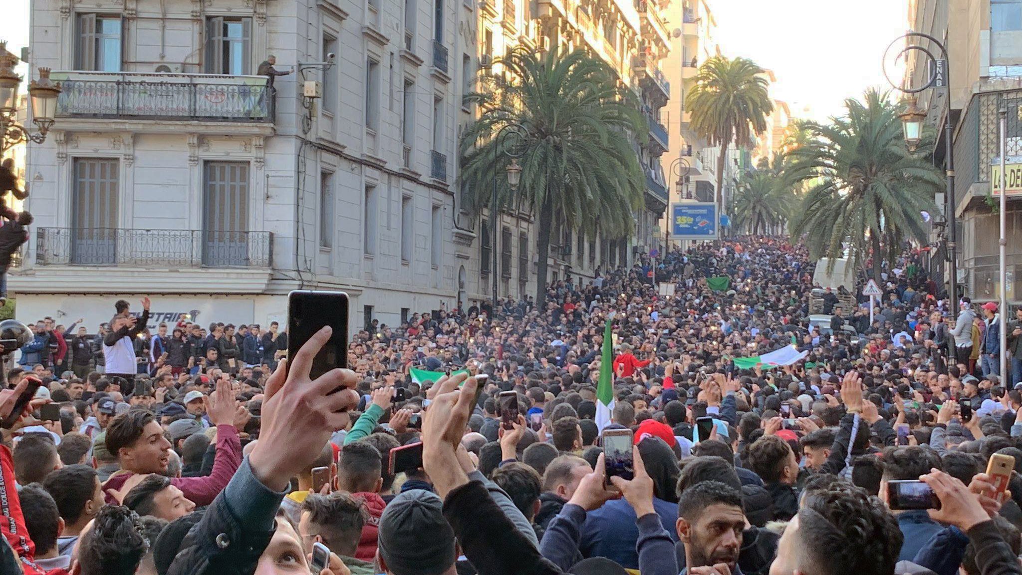 L'image montre une grande foule de personnes rassemblées dans une rue. Les participants semblent manifester, brandissant des drapeaux et levant des mains, certains tenant des téléphones pour filmer. On peut voir des bâtiments en arrière-plan, ainsi que des palmiers qui ajoutent une touche estivale à la scène. L'atmosphère semble dynamique et engagée, illustrant un moment de solidarité ou de protestation.