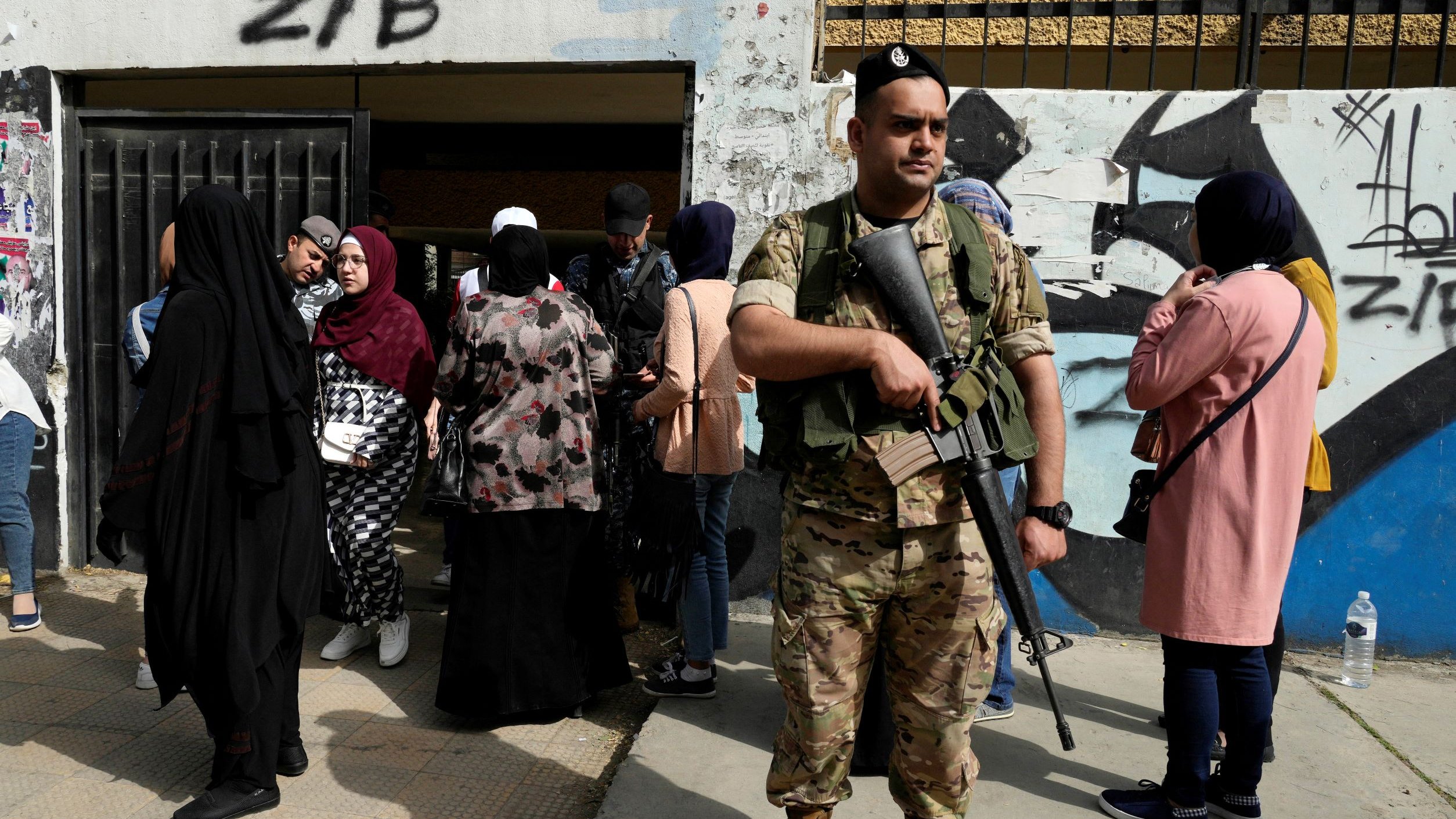L'image montre un soldat armé en tenue militaire, se tenant debout et vigilant à l'extérieur d'un bâtiment. Au fond, plusieurs femmes sont rassemblées, certaines portant des hijabs et des vêtements de différentes couleurs. Le cadre semble urbain, avec des murs décorés de graffitis. L'atmosphère semble chargée de tension, avec la présence militaire suggérant un contexte de sécurité ou de contrôle.