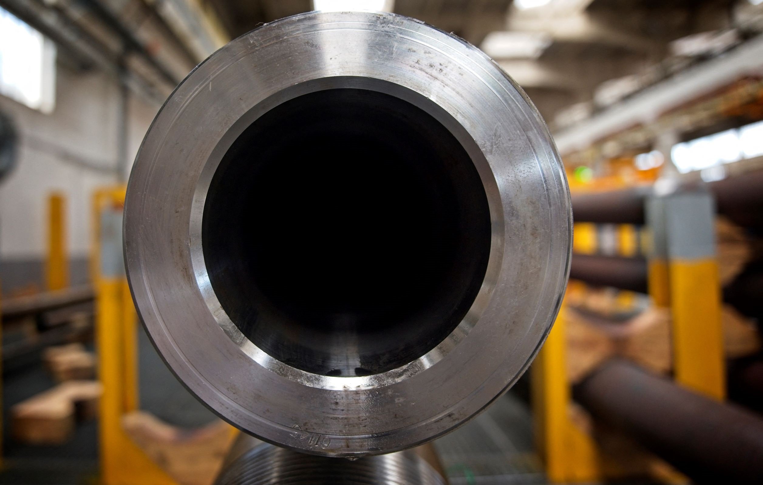 L'image montre la vue de l'intérieur d'un grand tube métallique. On peut voir la texture lisse et brillante du métal au niveau de l'ouverture, tandis que l'intérieur du tube est sombre. En arrière-plan, des éléments d'un environnement industriel sont visibles, avec d'autres tubes et structures, suggérant un lieu de fabrication ou d'assemblage. L'éclairage est principalement naturel, créant un effet contrasté sur la surface du tube.