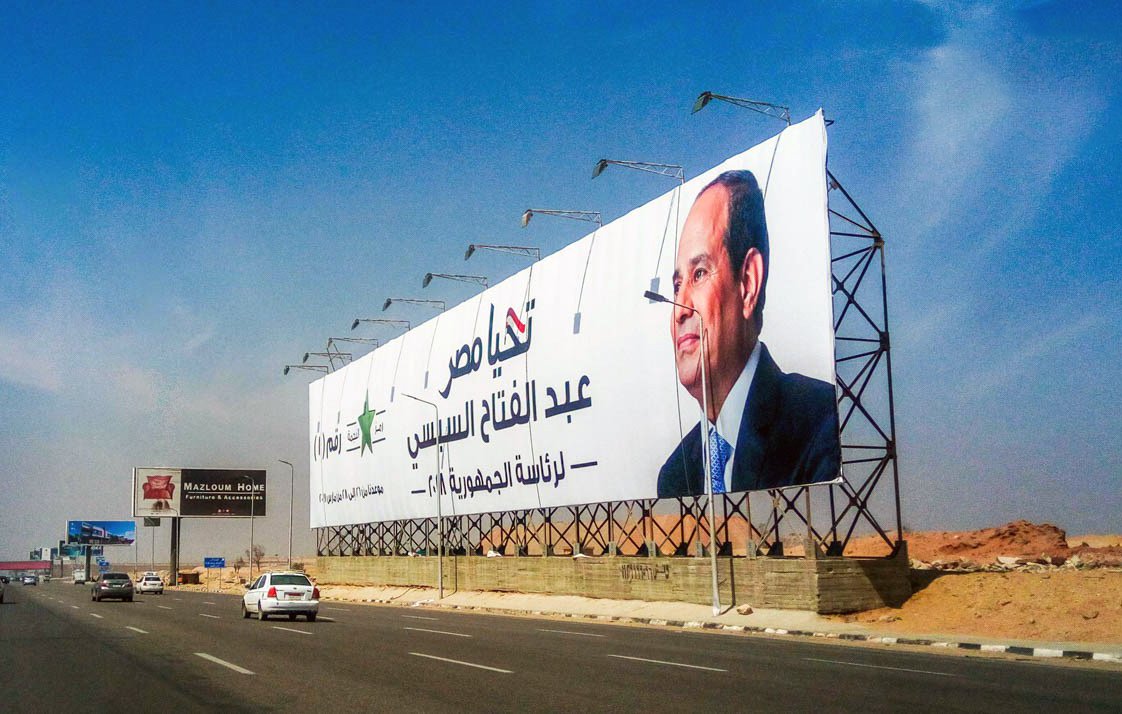 L'image montre un grand panneau publicitaire situé le long d'une route. Sur ce panneau, il y a une photo d'un homme en costume, accompagné d'un texte en arabe. La scène est prise dans un environnement urbain, avec une route et des véhicules en mouvement. Le ciel est clair et présente des teintes de bleu. Le panneau semble promouvoir une personnalité politique ou un événement lié à celle-ci.