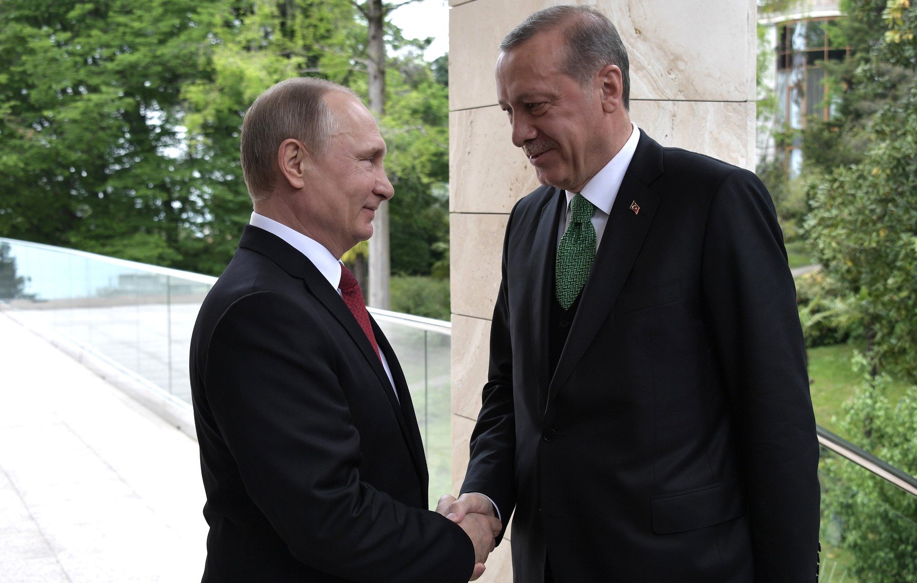 L'image montre deux hommes en costume, se serrant la main. L'un d'eux porte un costume noir avec une cravate rouge, tandis que l'autre est également en costume noir, mais avec une cravate verte. Ils se tiennent à l'extérieur, devant un fond de verdure et d'architecture moderne. L'atmosphère semble conviviale et formelle.