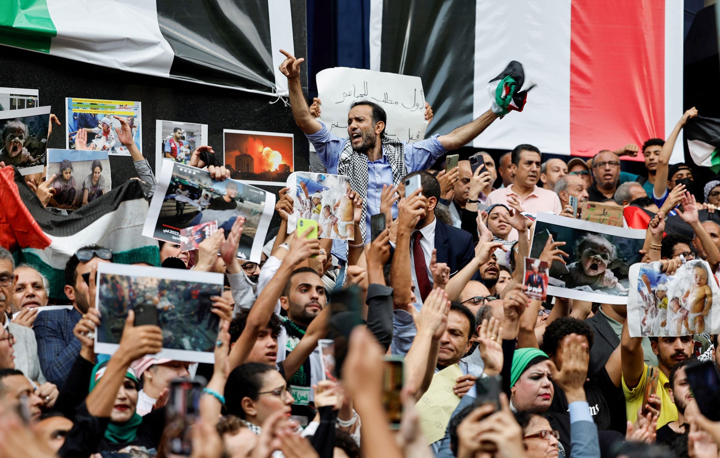 L'image montre une foule rassemblée lors d'une manifestation, avec des personnes brandissant des pancartes et des photos. Au centre, un homme lève les bras en signe de victoire ou de protestation. La foule exprime visiblement des émotions fortes, que ce soit la colère ou la solidarité. Des drapeaux sont également présents dans le décor, ajoutant à l'intensité de la scène. On ressent une atmosphère d'engagement et de mobilisation collective.