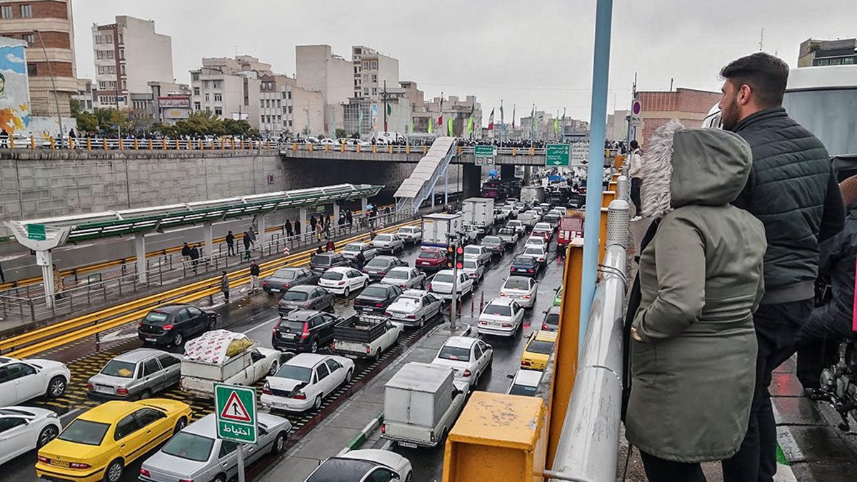L'image montre une scène de circulation dense dans une ville. On peut voir de nombreuses voitures de différentes marques immobilisées dans un embouteillage. À l'arrière-plan, on distingue des bâtiments urbains et une route à plusieurs niveaux. Il y a des panneaux de signalisation visibles, et deux personnes se tiennent sur un balcon ou une balustrade, observant la circulation en contrebas. Le ciel est nuageux, ce qui suggère un temps gris et éventuellement pluvieux.