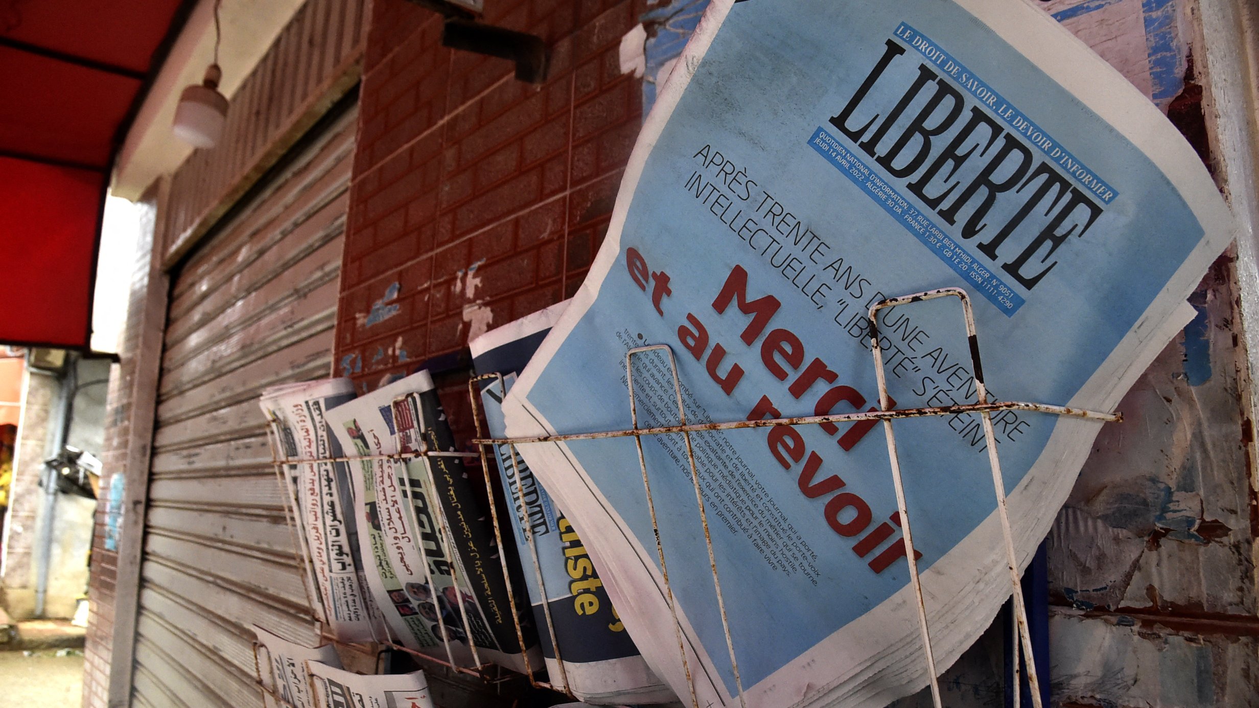 L'image montre un présentoir de journaux, où plusieurs titres sont visibles. Le journal en premier plan affiche le mot "LIBERTÉ" en lettres majuscules, accompagnés d'un sous-titre qui semble évoquer un sujet d'actualité. Les autres journaux à côté sont disposés de manière ordonnée, avec des titres variés, ce qui suggère un point de vente de presse dans un environnement urbain. Le mur en arrière-plan, ainsi que le toît rouge au-dessus, ajoutent à l'atmosphère locale de la scène.