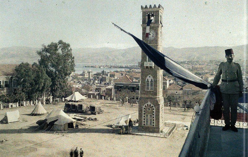L'image montre une vue panoramique d'une place animée avec un grand bâtiment en pierre qui pourrait être une horloge ou une tour emblématique. À proximité, un soldat en uniforme se tient près de la balustrade, observant la scène. On peut voir des tentes disposées sur la place, peut-être pour un camp militaire ou une exposition. En arrière-plan, on distingue des collines et un plan d'eau, ajoutant à la profondeur du paysage. Le ciel semble dégagé, créant une atmosphère paisible.