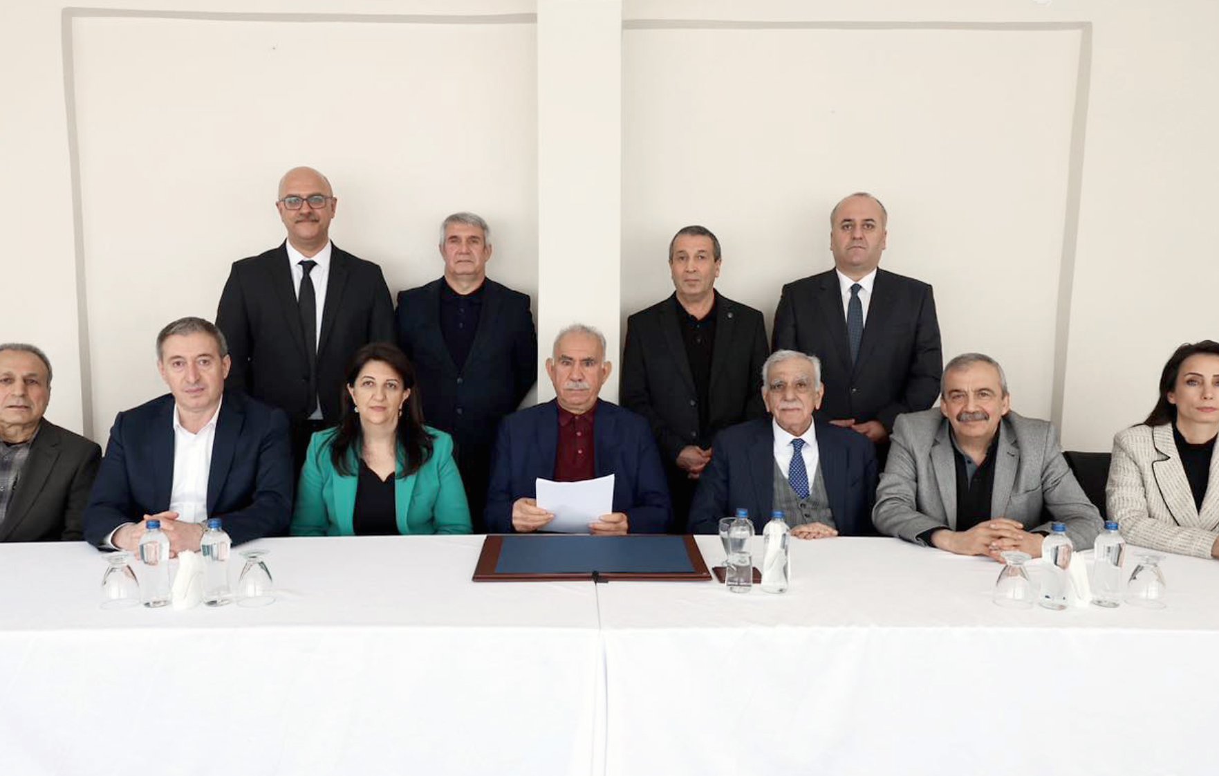 L'image montre un groupe de douze personnes assises derrière une table blanche. Il y a des hommes et des femmes, tous vêtus de costumes et de tenues professionnelles. L'ambiance semble formelle, et une personne au centre tient un document.