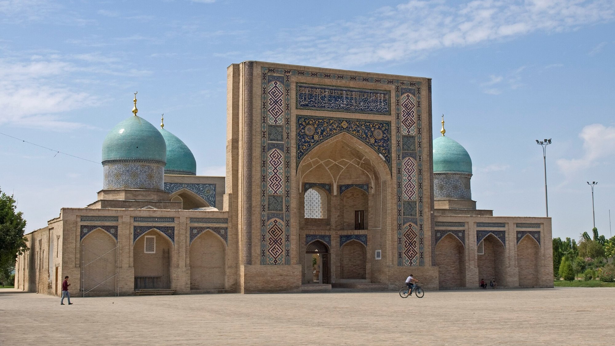 Bâtiment en brique avec de grandes arches et coupoles turquoise sous un ciel bleu.
