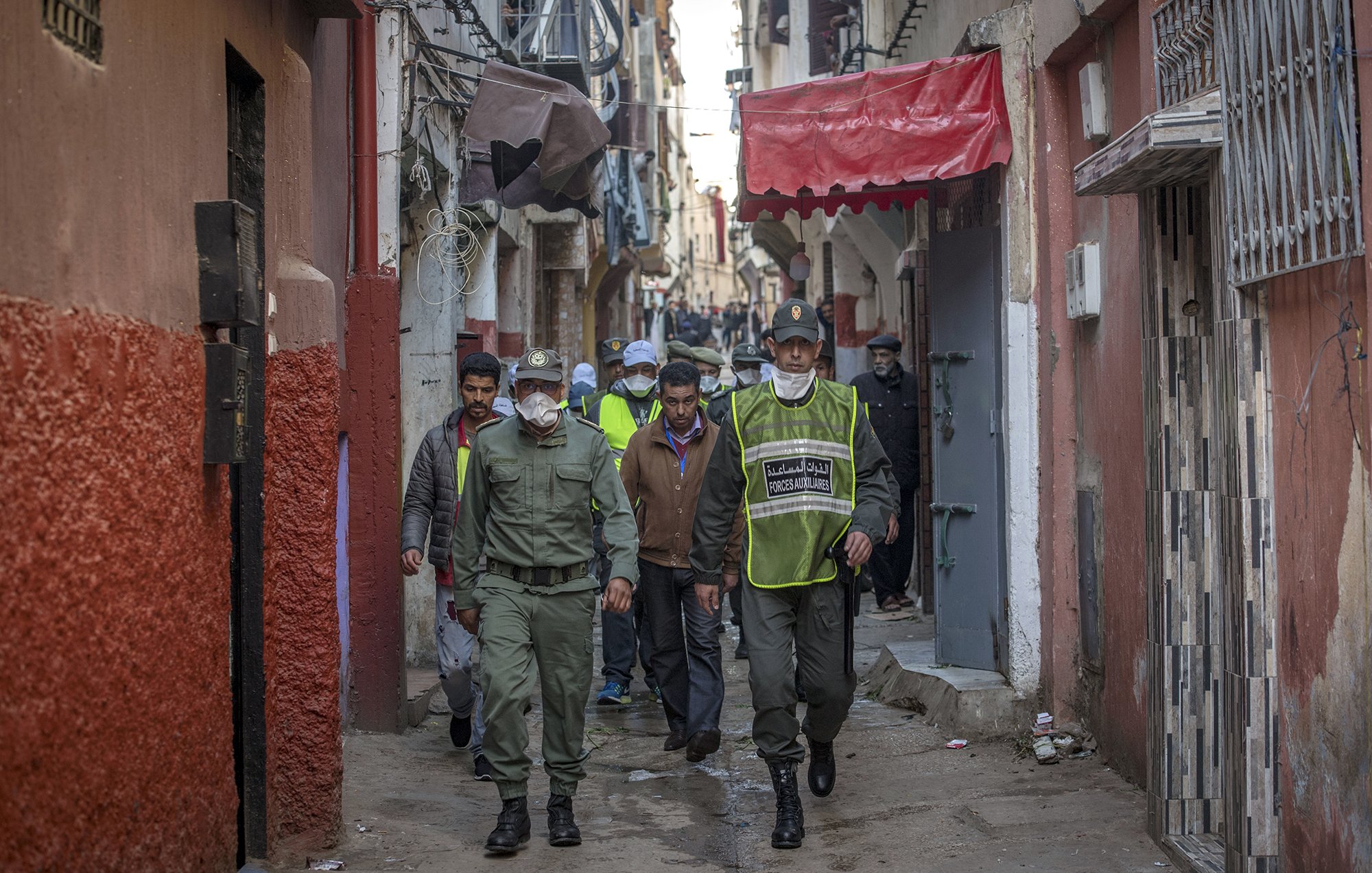 L'image montre une ruelle étroite et animée, typique d'une ville marocaine. Au premier plan, des agents de police ou des membres de la sécurité patrouillent. Ils portent des uniformes et des masques, indiquant une attention à la sécurité ou à la santé publique. En arrière-plan, on peut voir des habitants et d'autres personnes dans la rue, ainsi que des bâtiments aux façades colorées. L'atmosphère semble à la fois dynamique et surveillée, reflétant la vie quotidienne dans ce type d'environnement urbain.