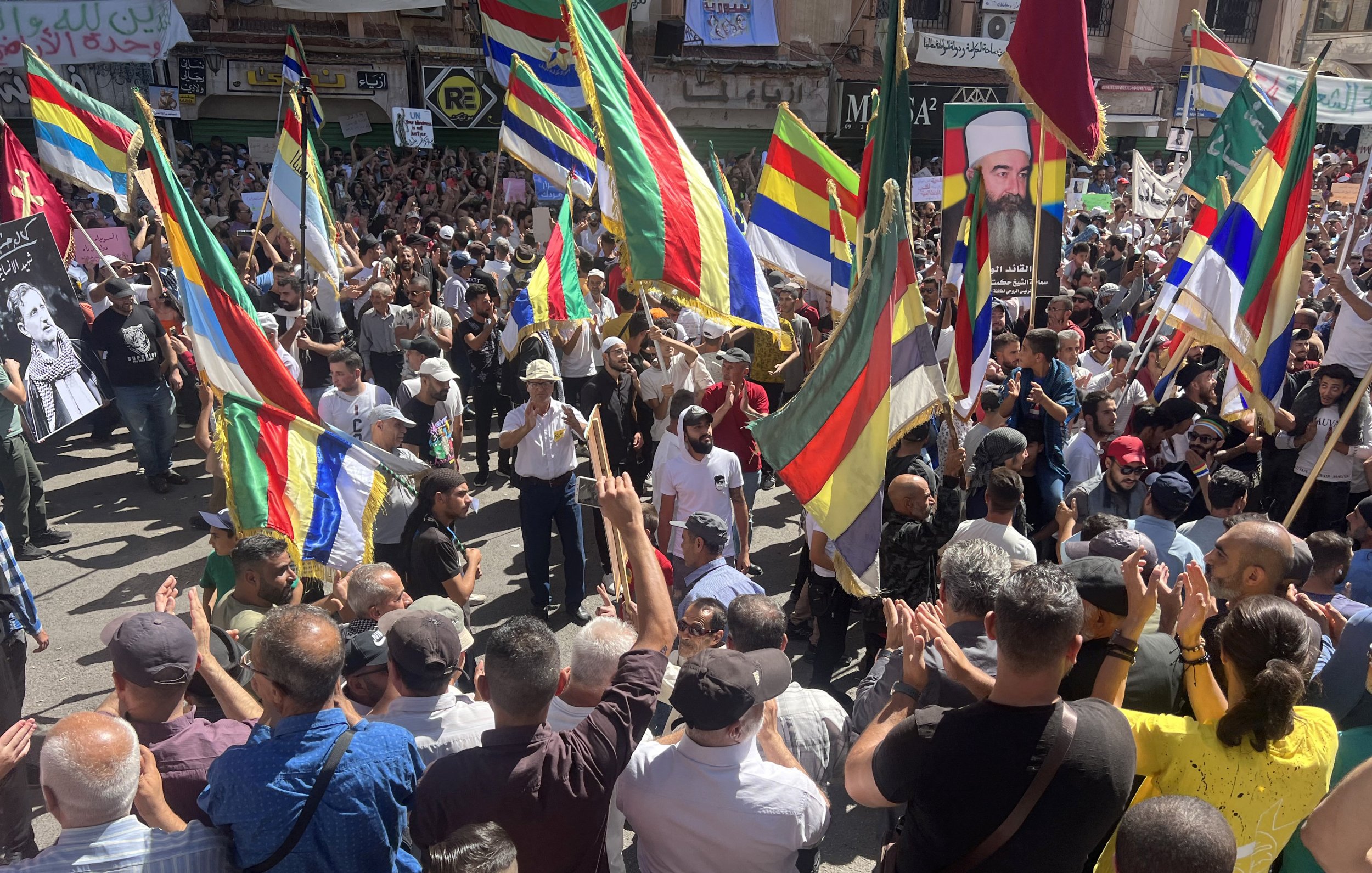 L'image montre une grande foule rassemblée lors d'une manifestation. Les participants tiennent des drapeaux colorés, probablement de différentes communautés ou groupes. On peut voir des visages de personnes connues affichés sur des pancartes. L'atmosphère semble dynamique et engagée, avec des manifestants applaudissant et exprimant leurs opinions. Le décor urbain suggère que l'événement se déroule dans une ville animée.