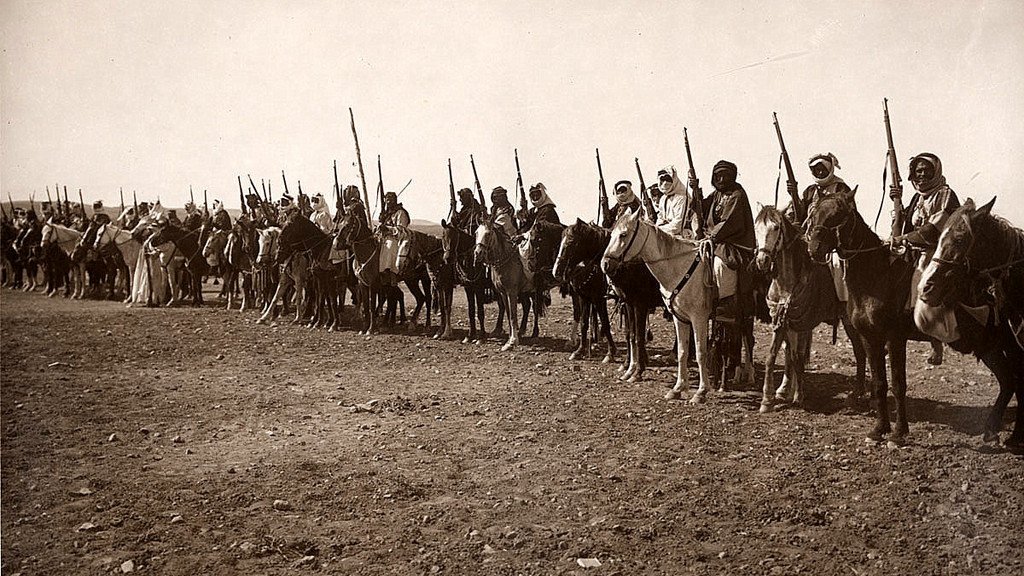 L'image montre une longue ligne de cavaliers en rang, tous montés sur des chevaux. Chaque cavalier est équipé d'une lance ou d'un bâton, et beaucoup portent des vêtements traditionnels. L'arrière-plan semble désertique, avec un sol rugueux et peu de végétation. L'atmosphère de l'image évoque une scène historique ou militaire, mettant en avant des guerriers ou des membres d'une tribu. Les cavaliers adoptent une posture fière et attentive, prêts pour une action ou une procession.