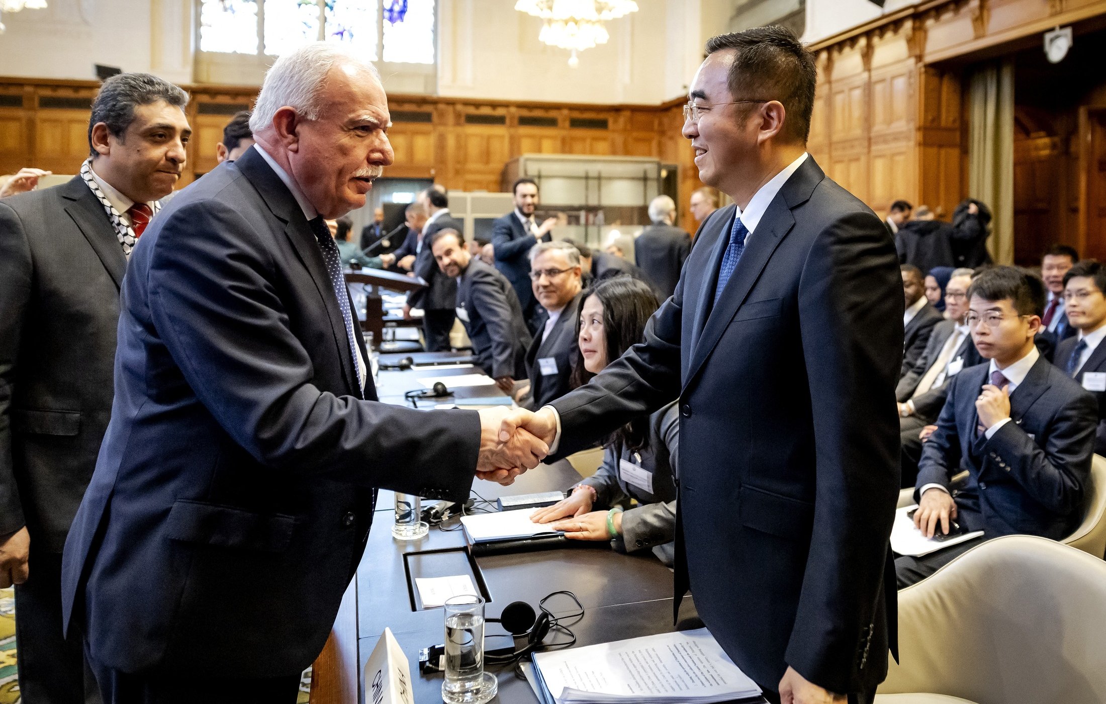 L'image montre une scène de réunion ou de conférence, probablement dans un cadre formel. Deux hommes se serrent la main, souriant, ce qui évoque une interaction chaleureuse ou un accord. En arrière-plan, on aperçoit d'autres participants qui semblent également engagés dans des discussions. La salle est spacieuse, avec une décoration élégante, des fenêtres laissant passer une lumière naturelle, et des sièges disposés autour d'une grande table. L'atmosphère semble professionnelle et collaborative.