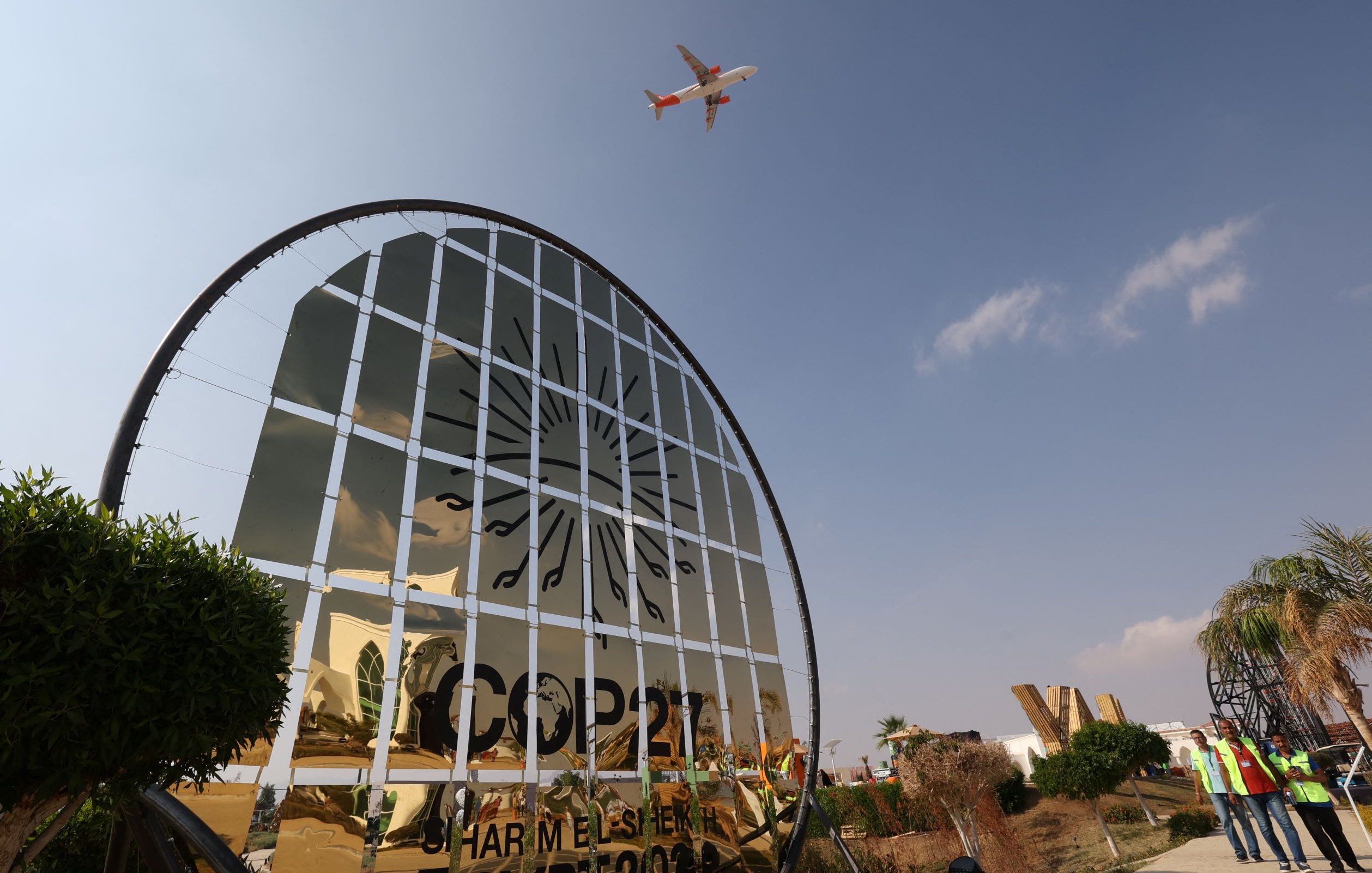 L'image montre un panneau de bienvenue à la COP27, ornée d'un design en miroir qui reflète le ciel. On aperçoit un avion volant au-dessus, suggérant une activité aérienne dans la région. Le paysage présente des éléments végétaux, comme des plantes et des arbres, et des personnes portant des vestes de sécurité sont visibles, indiquant une ambiance de préparation pour l'événement. Le ciel est clair avec quelques nuages.