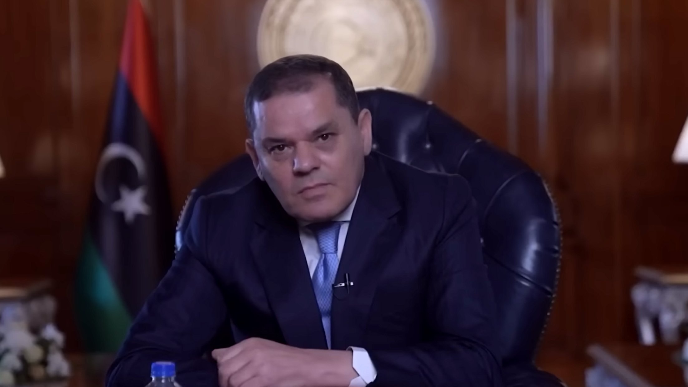 Homme en costume assis dans un bureau, drapeau libyen en arrière-plan.