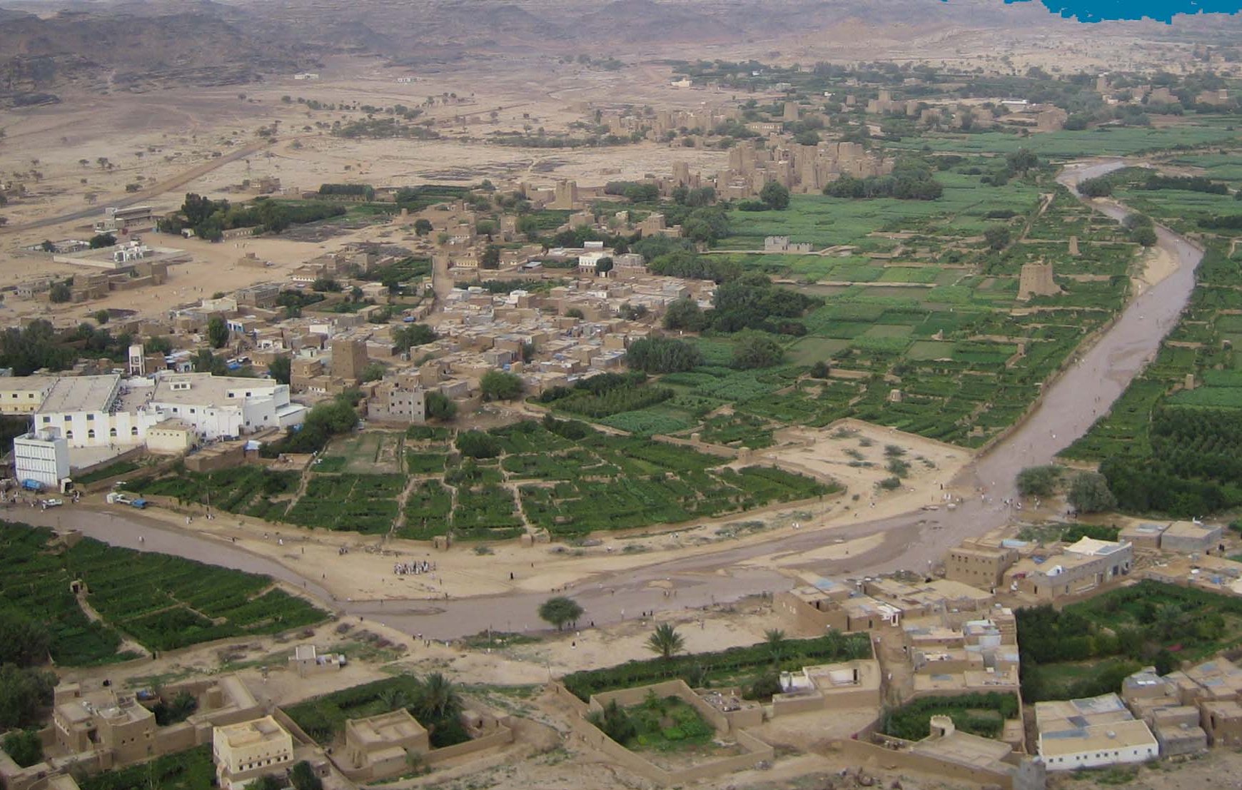 L'image montre un paysage rural, probablement une vallée verdoyante entourée de montagnes arides. Au premier plan, on peut voir des champs cultivés et des terres agricoles, formant un contraste avec les zones désertiques environnantes. Il y a également quelques maisons en terre ou en briques qui semblent faire partie d'un village. La rivière qui serpente à travers le paysage ajoute à la beauté de la scène, symbolisant une source vital d'eau dans cette région dry. Des traces de chemins et des gens peuvent également être aperçus, suggérant une vie active dans cette zone rurale.