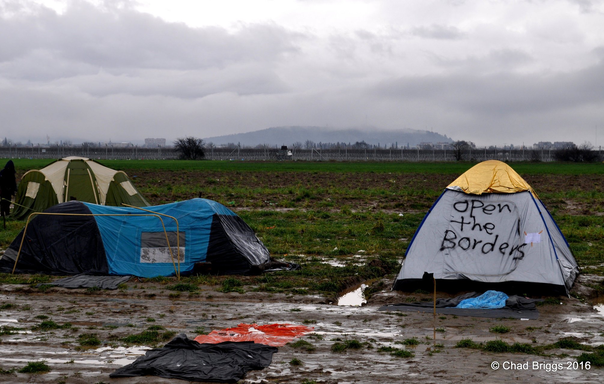 L'image montre un campement de tentes installé dans un terrain boueux. On aperçoit plusieurs tentes, dont l'une a un message écrit : "Open the Borders". Le paysage environnant est composé de champs et d'une arrière-plan nuageux, ce qui crée une atmosphère morose. Le sol est détrempé, indiquant probablement des conditions météorologiques difficiles.