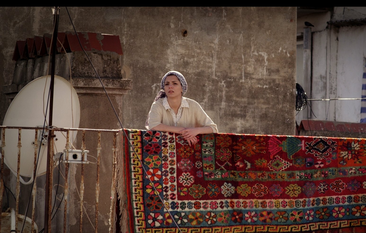 L'image montre une femme se tenant sur un balcon, regardant au loin. Elle porte un haut clair et un foulard sur la tête. Derrière elle, un grand tapis coloré est accrochée à la balustrade, avec des motifs détaillés et vibrants. En arrière-plan, on aperçoit un mur et une antenne satellite, ce qui suggère un environnement urbain. L'éclairage donne une atmosphère chaleureuse à la scène.