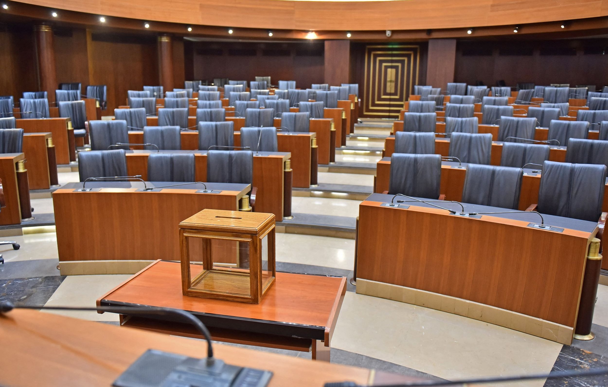 L'image montre une salle de réunion ou un parlement. Les sièges sont en bois, avec des dossiers noirs et sont disposés en rangées organisées. Au centre, il y a une petite table en bois avec une boîte en verre posée dessus. L'éclairage est doux, mettant en valeur les finitions en bois des murs et des meubles. L'ensemble de la salle dégage une atmosphère formelle et austère.