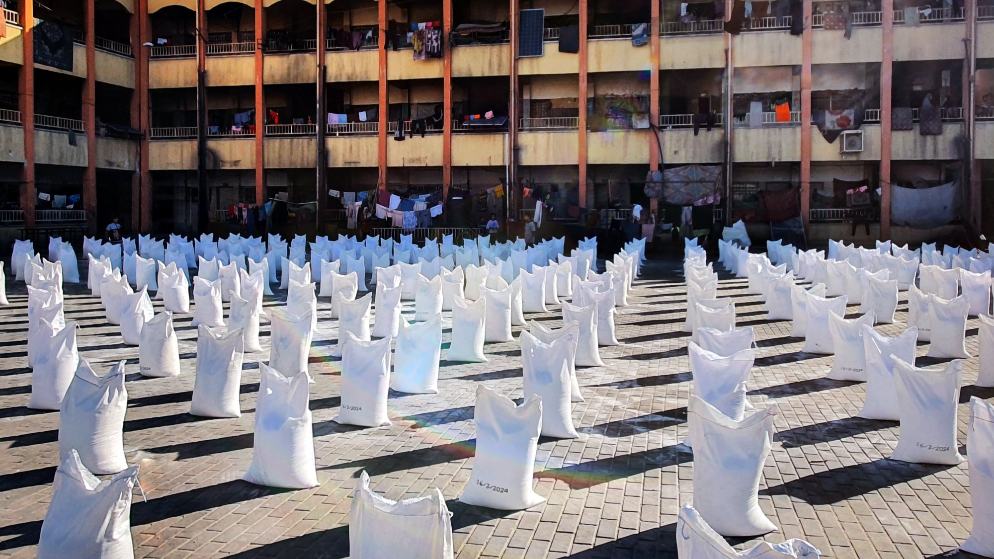 L'image représente un espace ouvert où de nombreux sacs blancs sont disposés en lignes. Ces sacs semblent être remplis de quelque chose, et le sol est constitué de pavés. En arrière-plan, on peut voir un bâtiment à plusieurs étages avec des balcons. Des vêtements sont suspendus à l'extérieur des balcons, suggérant une utilisation résidentielle de cet espace. La lumière du soleil éclaire la scène, créant un contraste entre les sacs blancs et l'environnement.