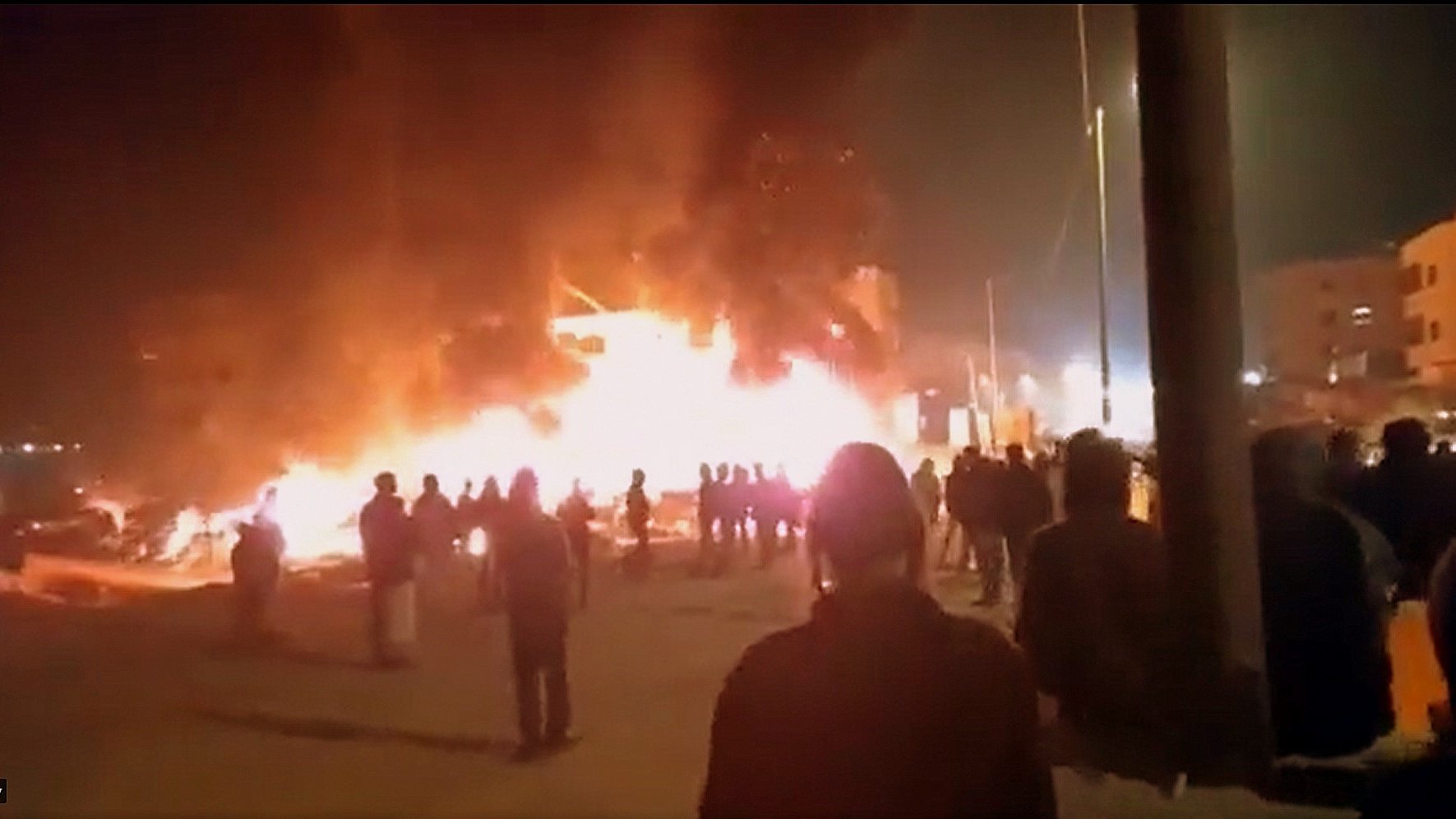L'image montre une scène nocturne où un bâtiment est enflammé, créant une grande colonne de feu et de fumée. Des personnes se tiennent au premier plan, observant la situation. On peut noter une ambiance de tension, avec des lumières des bâtiments environnants qui ajoutent à l'atmosphère dramatique. Les silhouettes des personnes sont découpées contre la lumière des flammes.