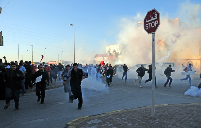 L'image montre une scène de manifestation. De nombreuses personnes courent dans une rue, entourées de fumée, probablement due à des gaz lacrymogènes. Au premier plan, on peut voir un panneau de signalisation "STOP". L'atmosphère semble tendue, et les manifestants semblent fuir une situation chaotique. Les participants portent des vêtements variés et certains tiennent des drapeaux.