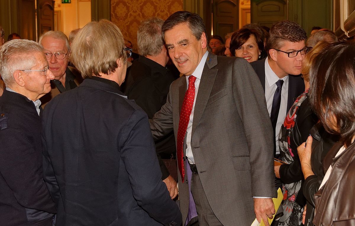 L'image montre un groupe de personnes réunies dans une salle, probablement lors d'un événement. Au centre, un homme en costume, avec une cravate à pois rouges, semble engager une conversation avec quelqu'un en lui serrant la main. D'autres personnes, en arrière-plan, observent ou participent à la discussion. L'ambiance semble conviviale, avec des sourires et des échanges entre les participants. Les murs sont décorés et ajoutent une touche d'élégance à la scène.