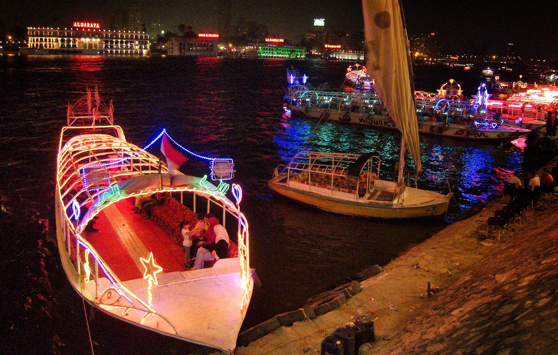 L'image montre un paysage nocturne animé le long d'une rivière. Plusieurs bateaux décorés de lumières colorées sont amarrés, créant une atmosphère festive. On peut voir des gens sur les bateaux et le long de la promenade, profitant de l'ambiance. En arrière-plan, des bâtiments illuminés ajoutent à la scène, avec des reflets dans l'eau. L'ensemble évoque une belle soirée en bord de rivière.