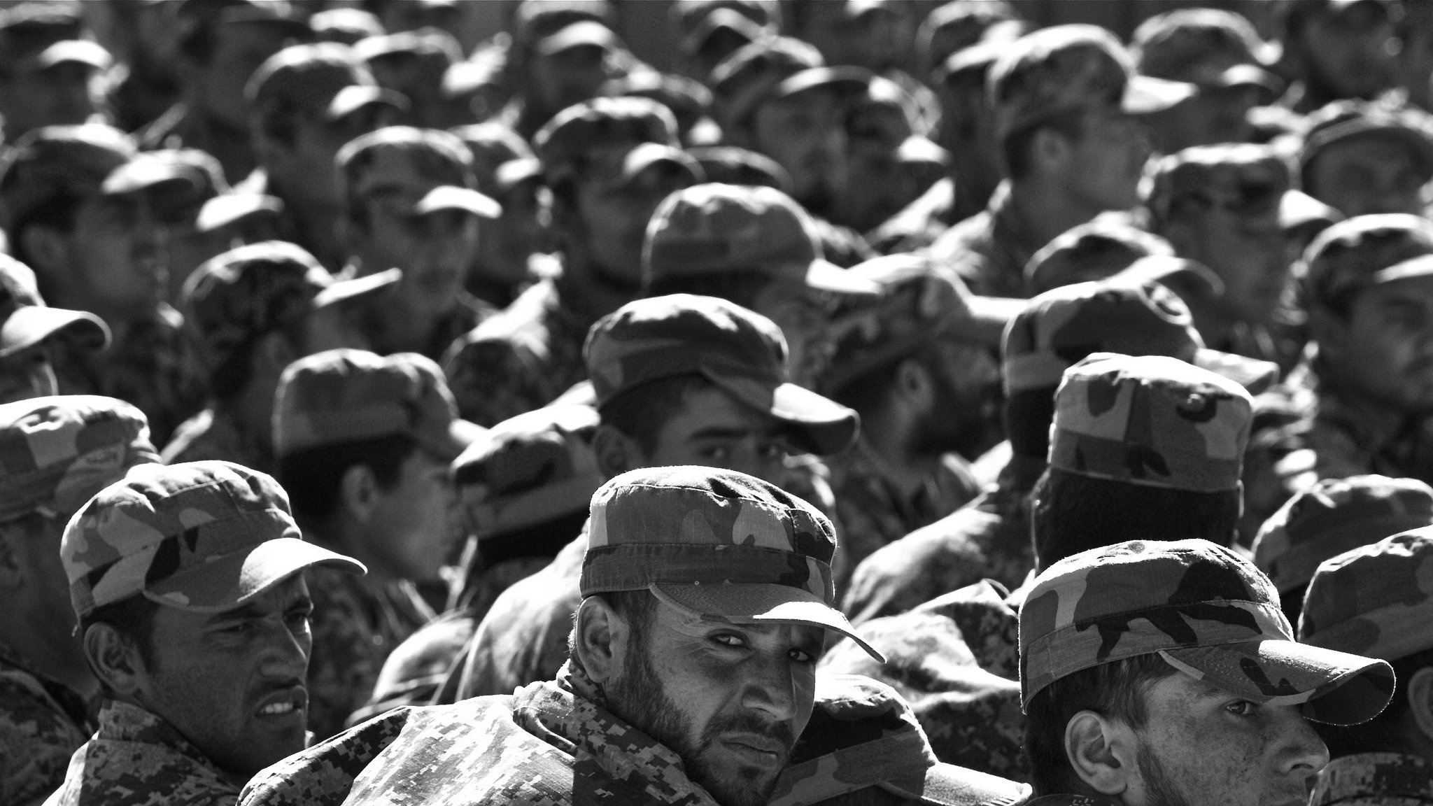 L'image montre un grand groupe de soldats en uniforme militaire, portant des casquettes assorties. Ils sont rassemblés et semblent attentifs, avec des expressions variées sur leurs visages. Le noir et blanc de la photo accentue les détails des uniformes camouflés et crée une atmosphère sérieuse, soulignant la discipline et la solidarité au sein du groupe.